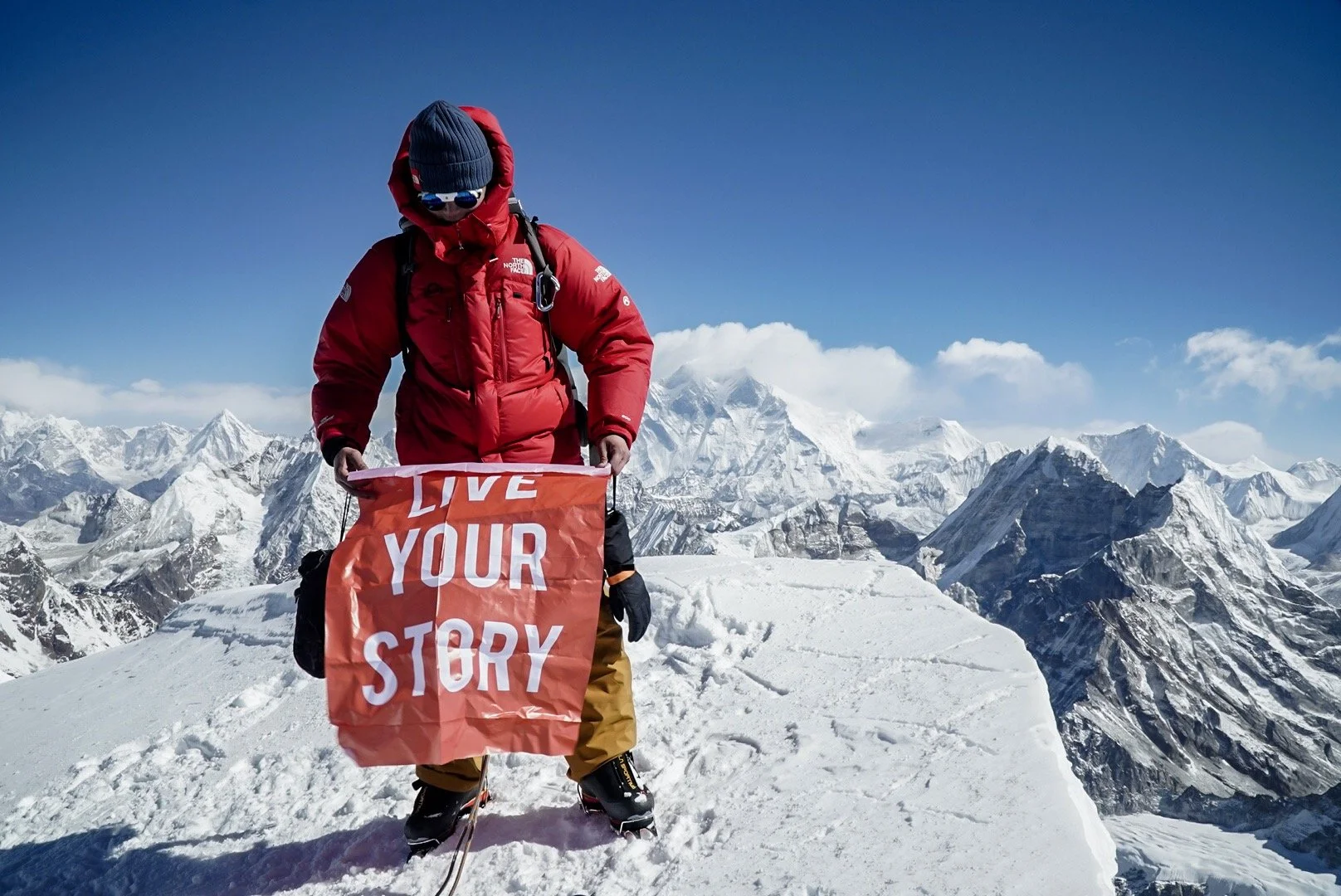 A mountain climber in red jacket and brown pants holding an orange banner that says 'LIVE YOUR STORY' on a snow-covered mountain peak with rugged snow-capped mountains in the background under a blue sky.