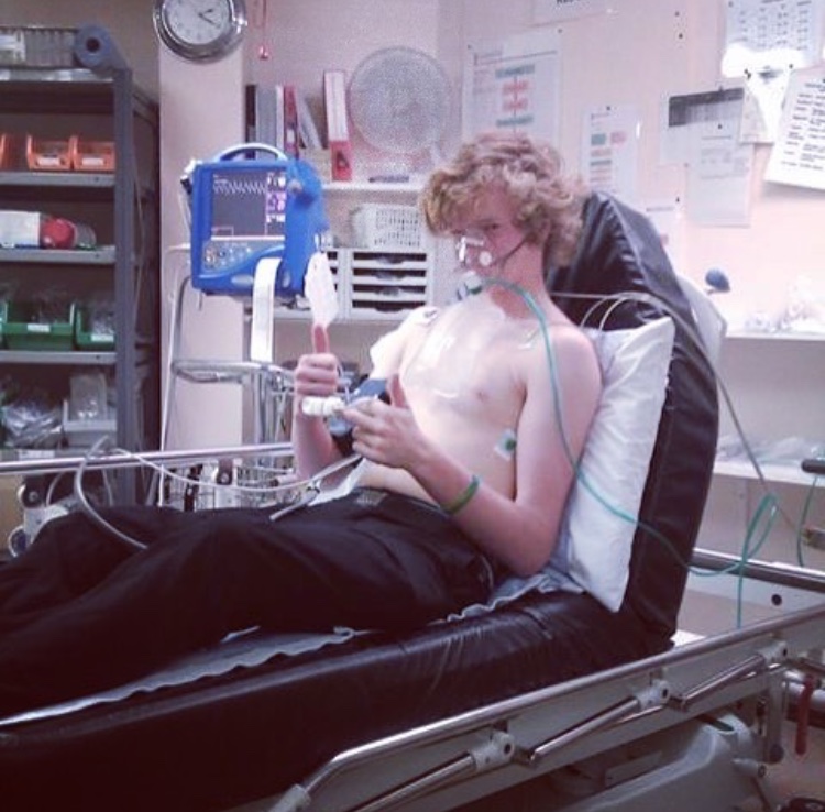 An image of Jago in a hospital bed, holding his thumbs up, wearing an oxygen mask