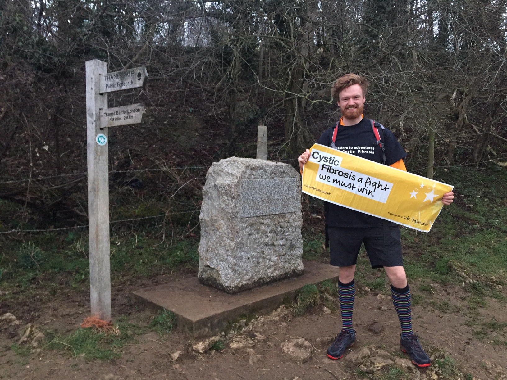 An image of Jago at the start of the Thames Path, holding a banner saying “Cystic Fibrosis- a fight we must win”