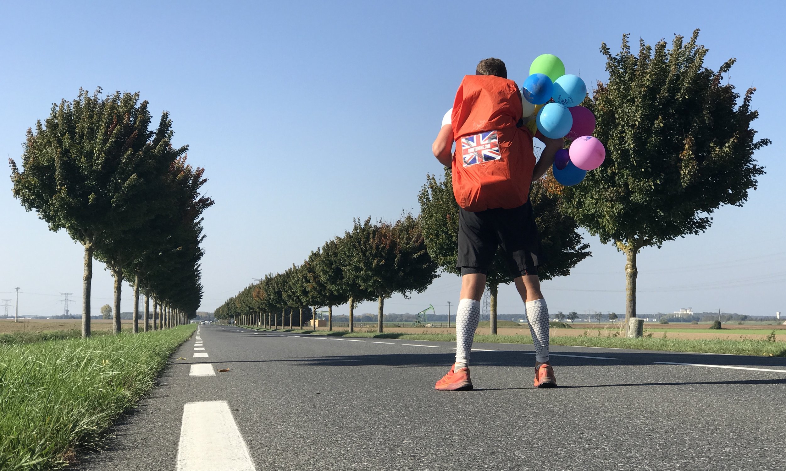 Image of Dan standing with his back to the camera, in a very straight road looking into the distance- wearing a bunch of colourful balloons on his back pack