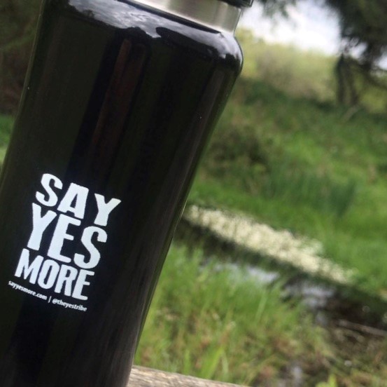 A black SayYesMore water bottle slants across half the photo, the other half is a blurry white and green spring landscape