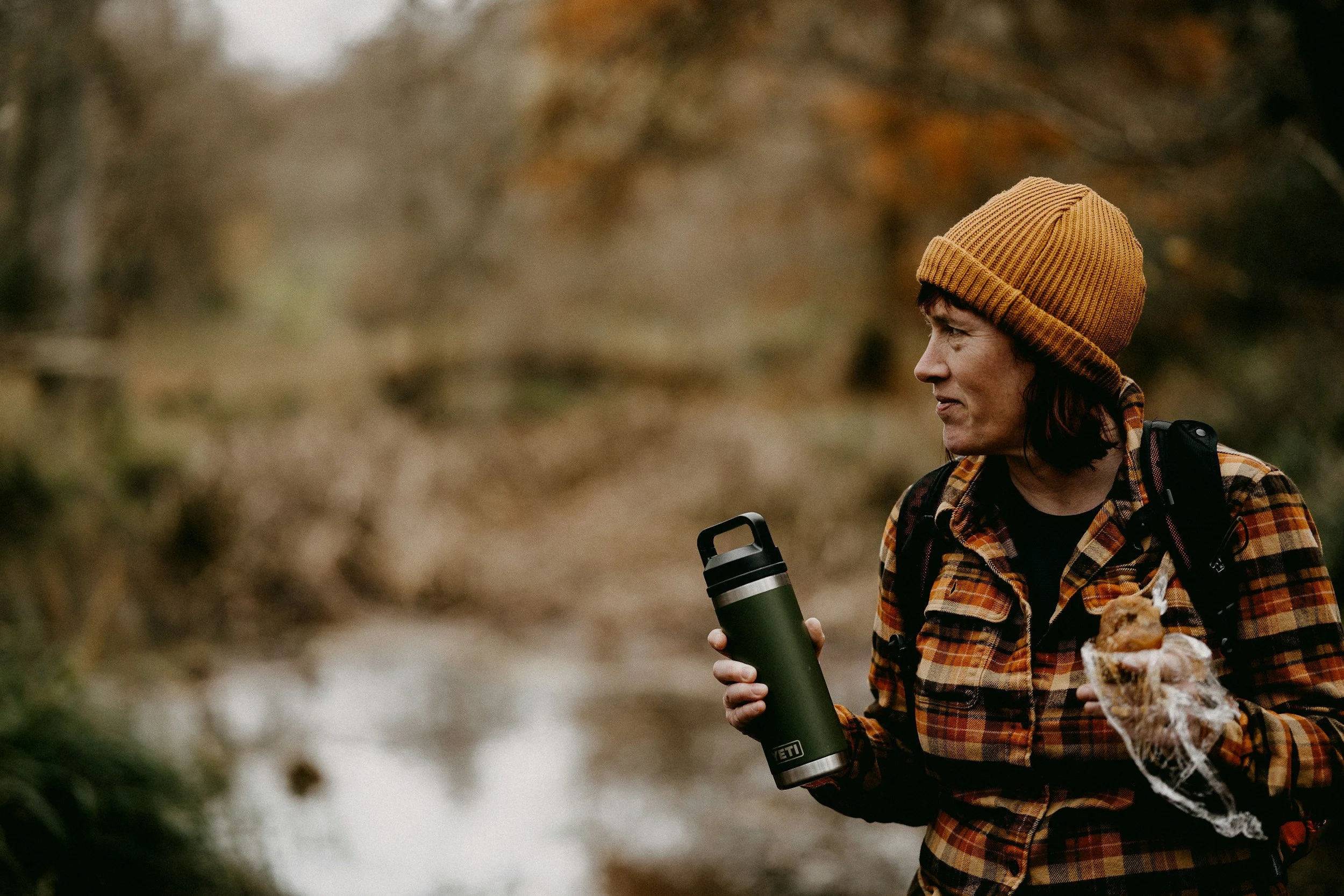 woman dressed in layers wearing a beanie, flannel, holding a water bottle, and a muffin while hiking