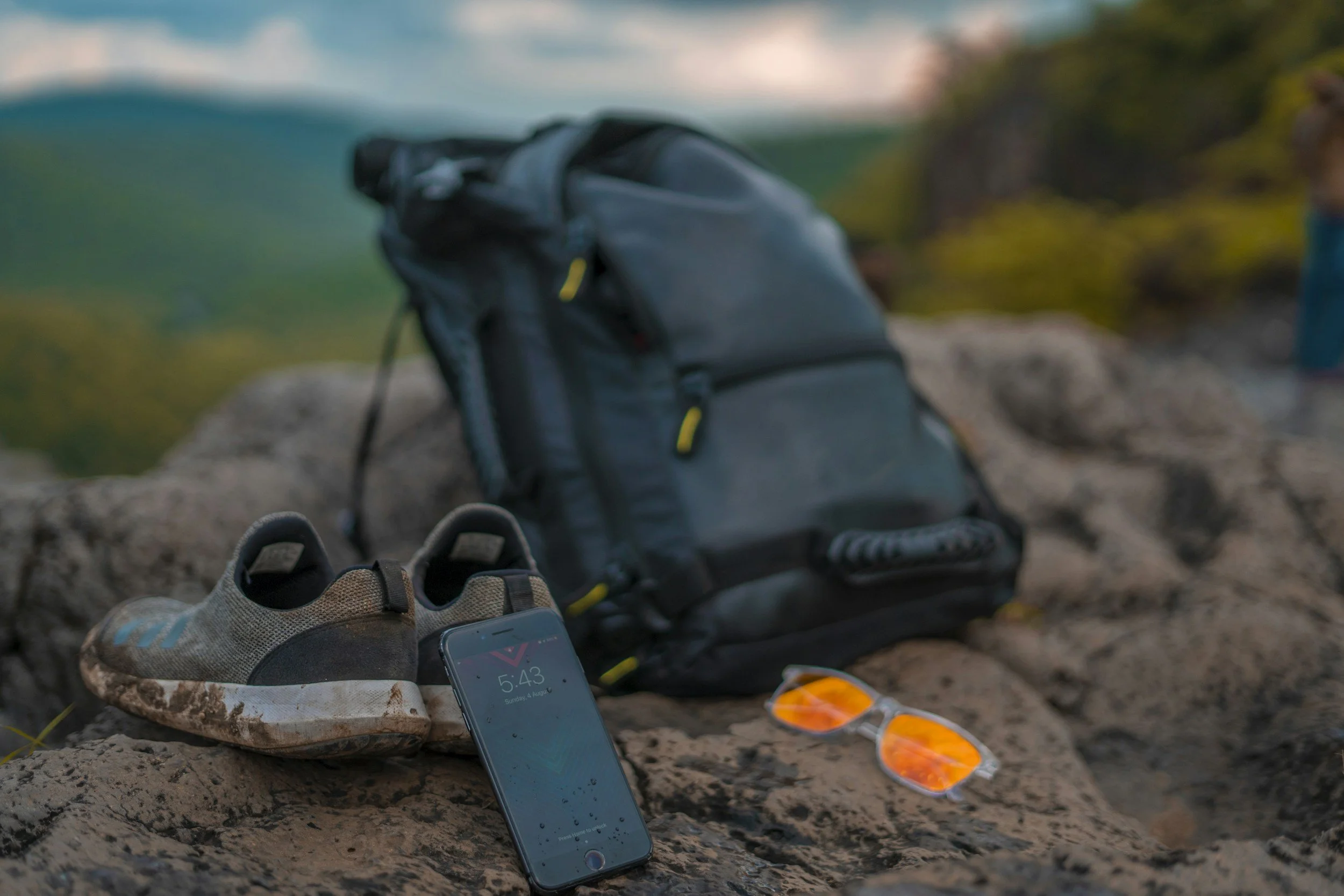 backpack, muddy shoes, phone, and sunglasses sitting on a rock in nature