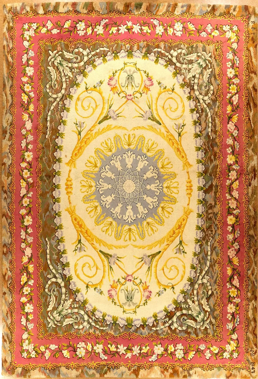 A decorative ornate rug with floral and scroll patterns, featuring a pink border with white flowers, a cream interior with yellow and gray design elements, and intricate floral motifs throughout.