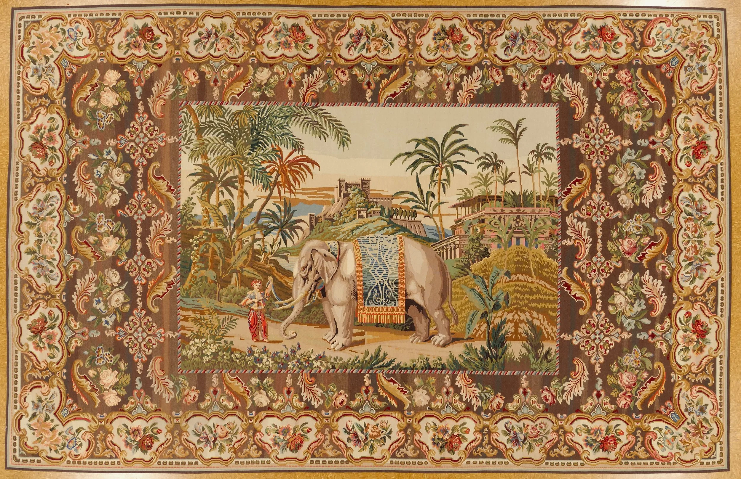 A decorative tapestry with a central scene of an elephant adorned with a colorful blanket, surrounded by palm trees and lush landscape, framed by intricate floral patterns on a brown background.