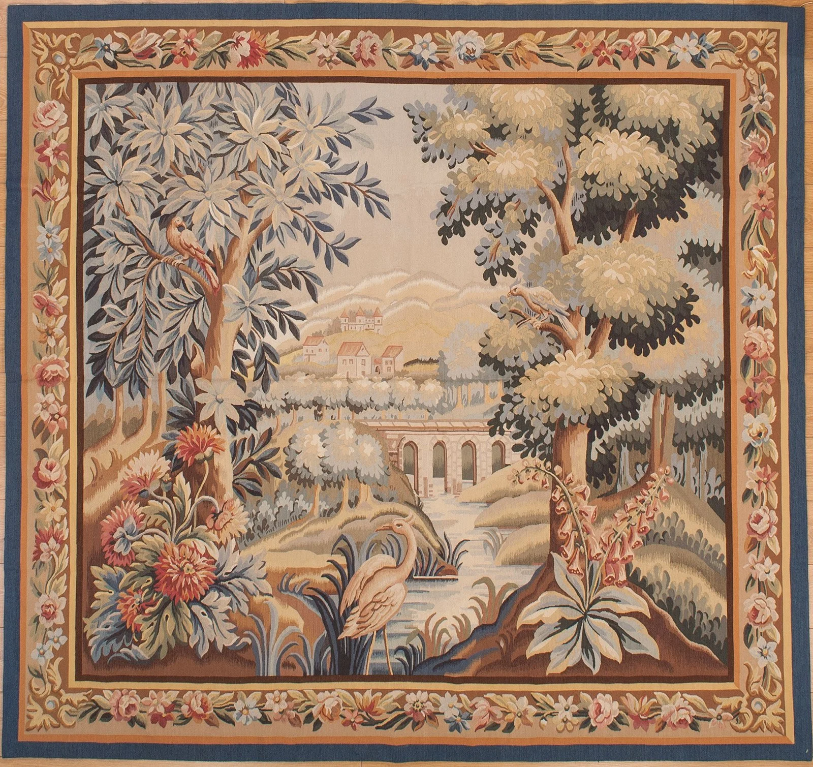 A detailed landscape tapestry depicting trees, birds, a river, a bridge, and houses in the background, surrounded by a floral border.