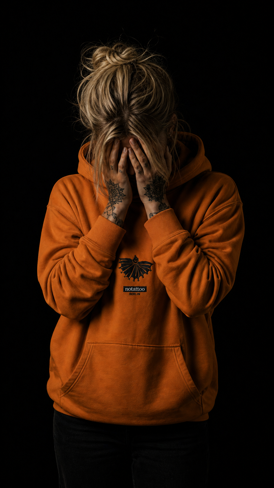 Front studio portrait of Adobe Yellow embroidered medieval bat hoodie by noSHBerlin, with face hidden by hands and tattooed fingers against a black background.