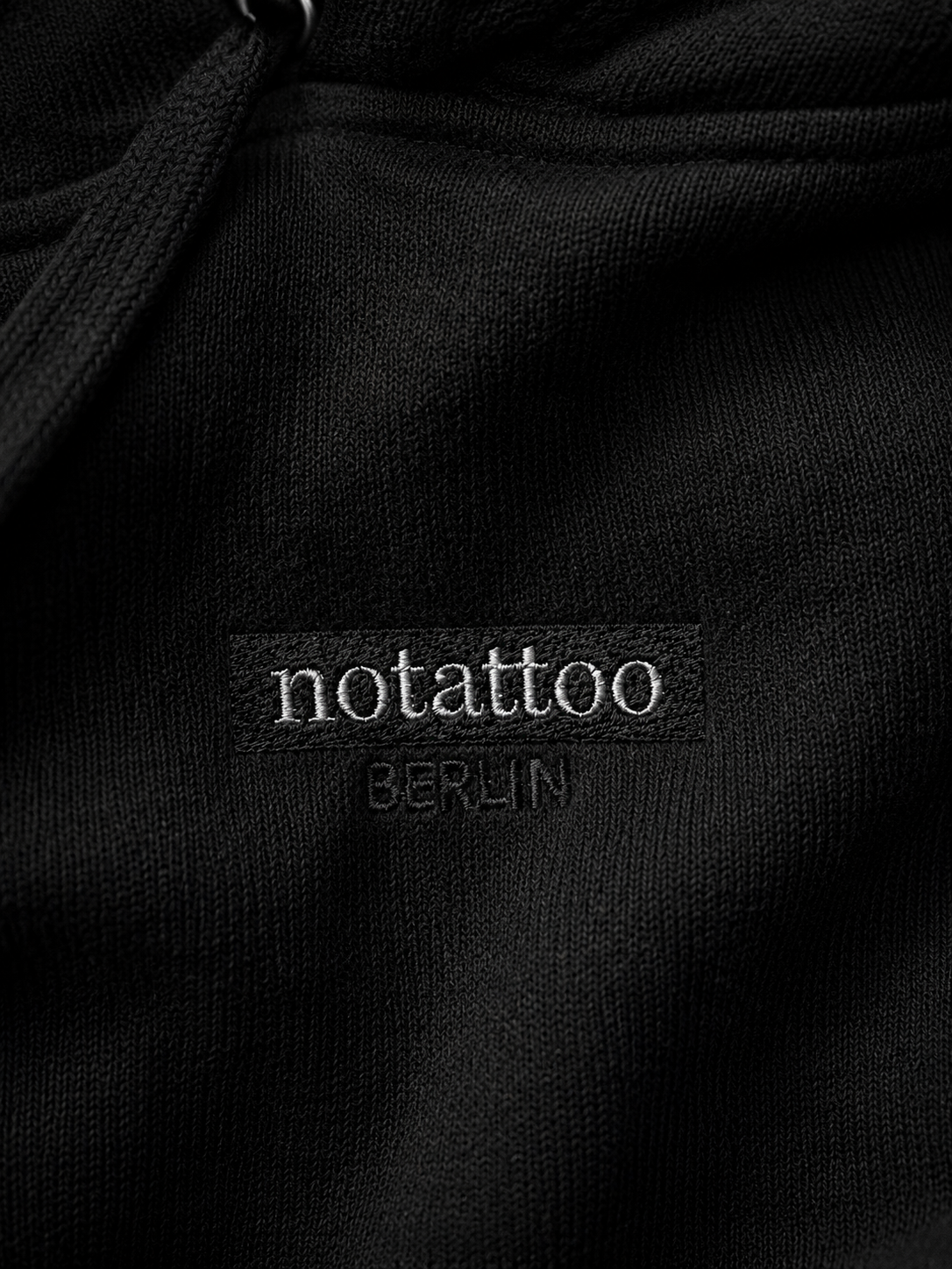 Close-up of embroidered noTATTOO Berlin logo on black hoodie fabric, showing the minimal everyday design of the hoodie.