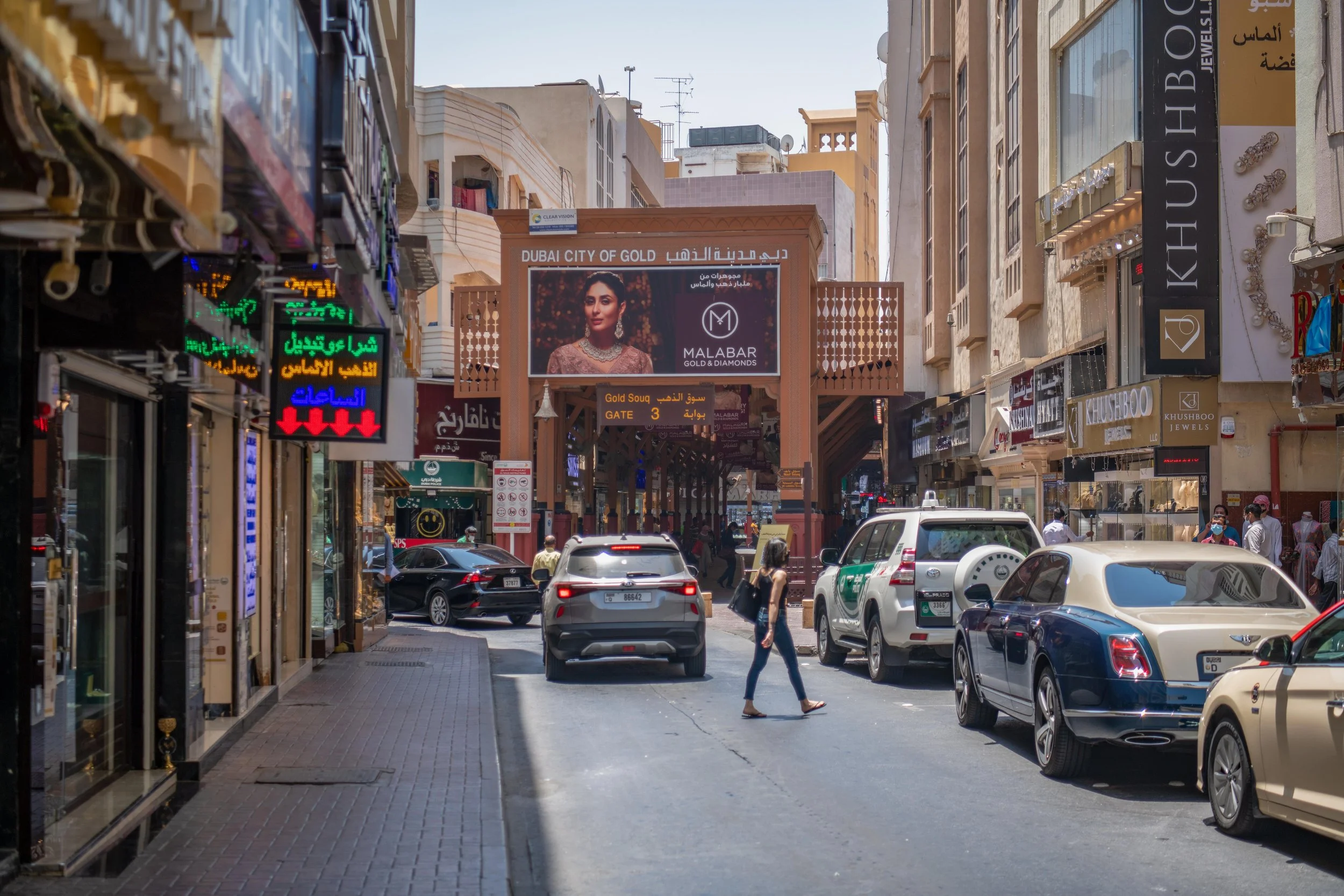 Woman walking through the bustling Gold Souk in Old Dubai, surrounded by traditional jewelry shops, taxis, and Arabic signage. The perfect intro to a cultural tour for parents visiting Dubai.