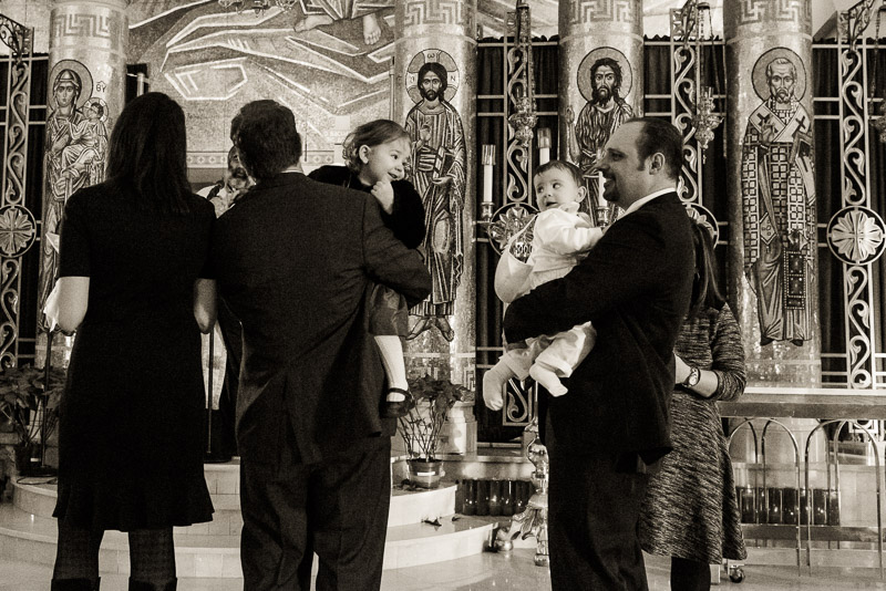 Two men each holding a child, the children looking at each other while the adults are listening to the priest.