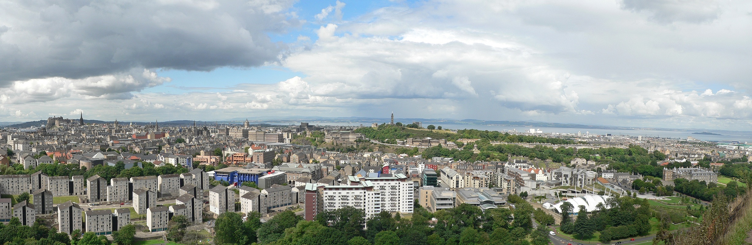 Spectacular panoramic image of Edinburgh and the Firth of Forth from Salisbury Crags. At left in the distance is Edinburgh Castle and the spire of the Hub, while the Nelson Monument stands tall atop Calton Hill just off the Center. Below it to the right are the Scottish Parliament and Holyrood Palace. Image Credit: Oliver-Bonjoch via Wikimedia Commons CC