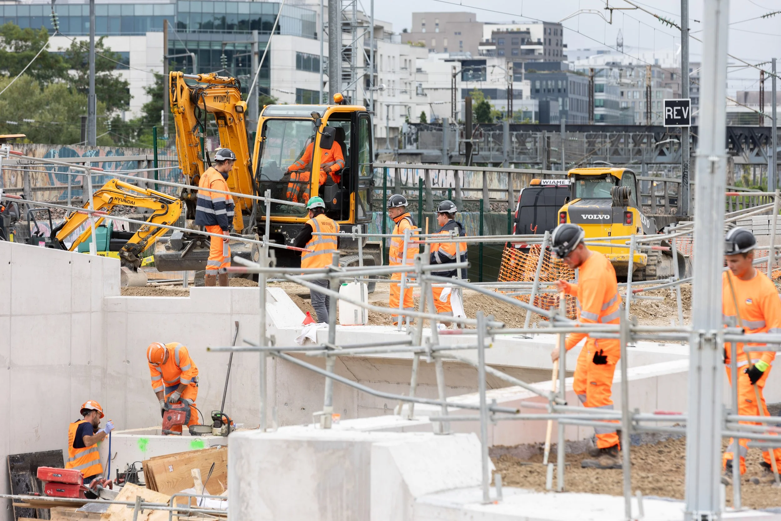 Des ouvriers travaillent sur un chantier de construction avec des engins de chantier et des barrières de sécurité, dans un environnement urbain.