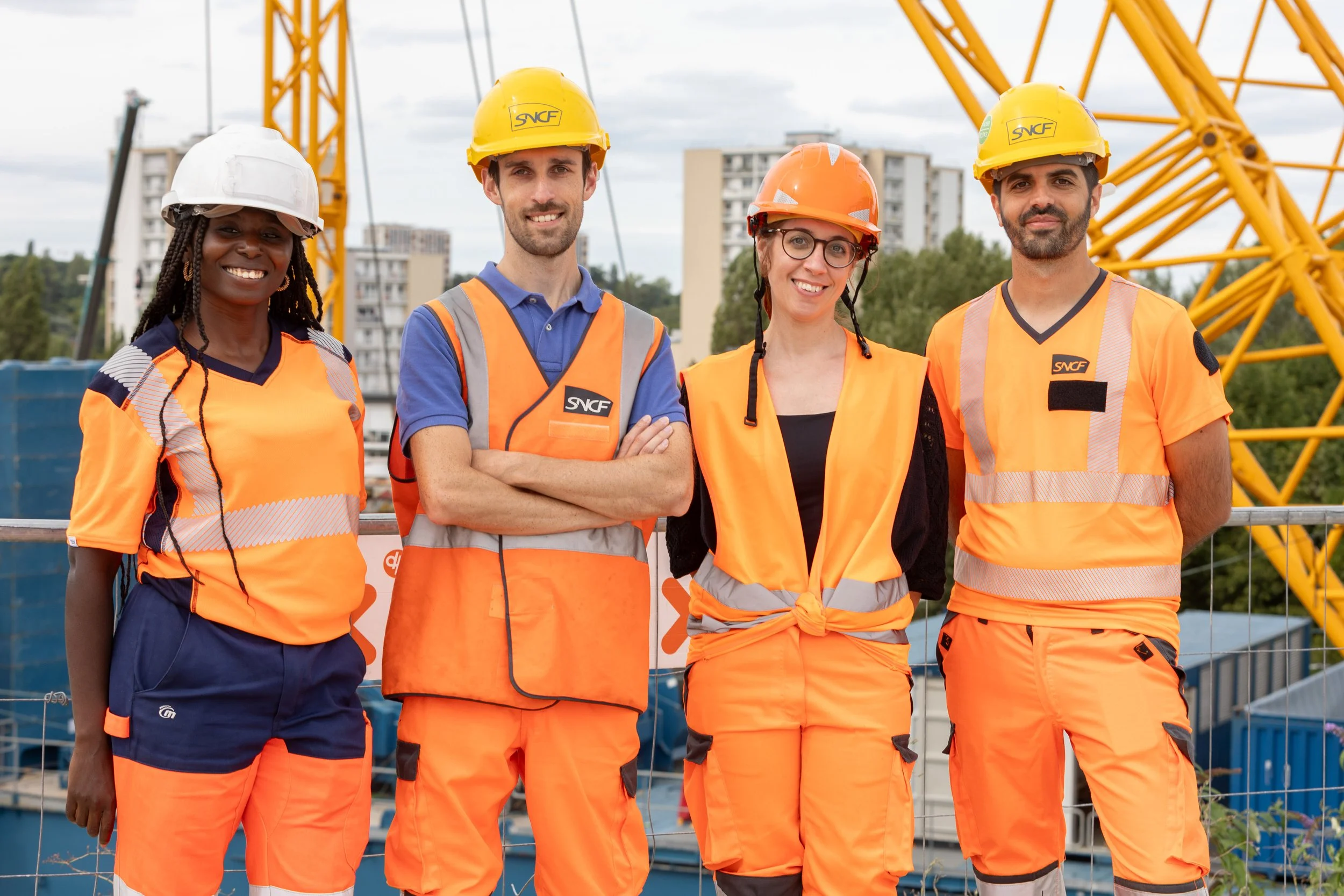 Quatre travailleurs portant des casques de sécurité et des vêtements de chantier orange, se tenant devant une structure métallique jaune sur un site de construction urbain.