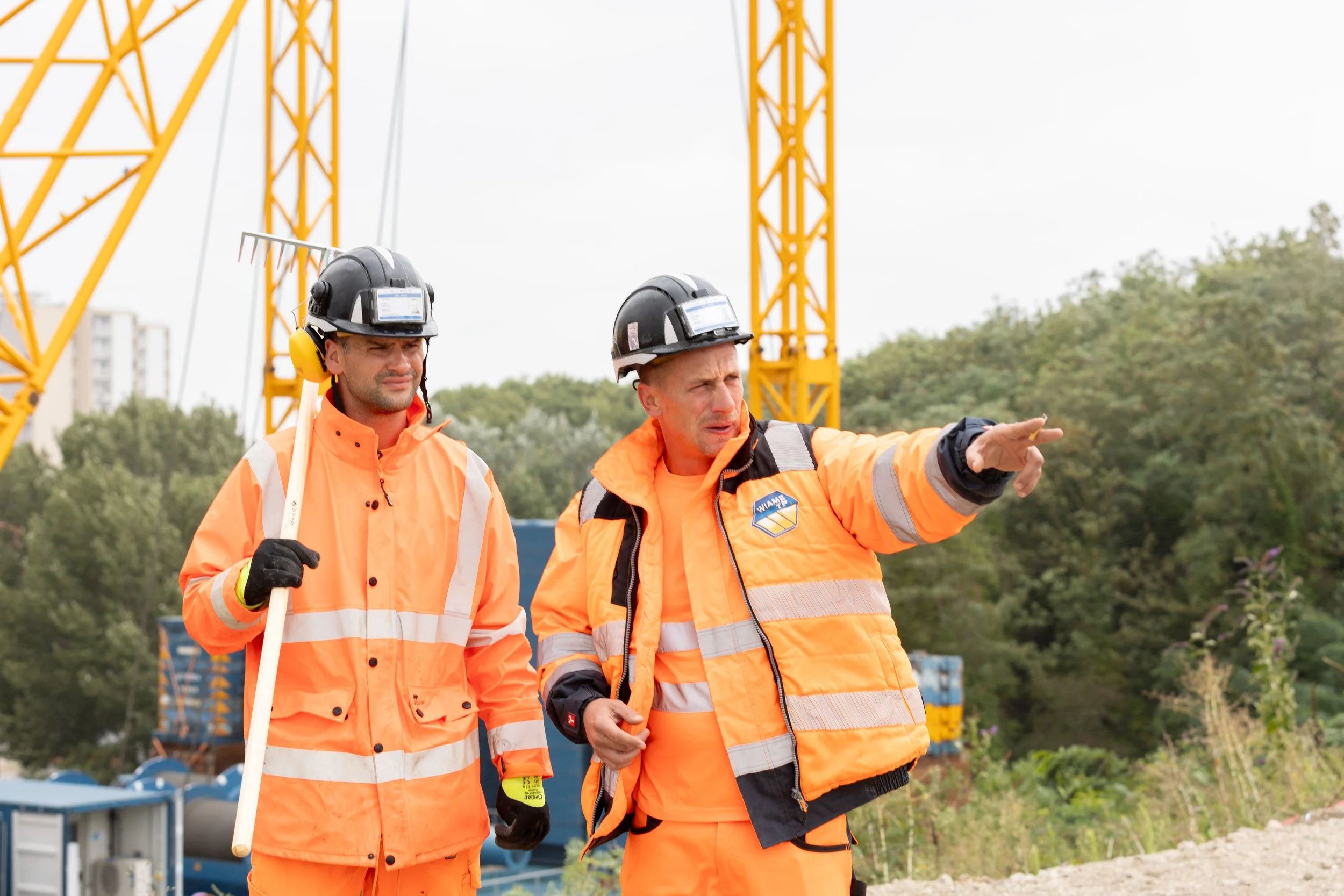 Deux hommes en uniforme de chantier orange discutent sur un site de construction avec une grue jaune en arrière-plan.