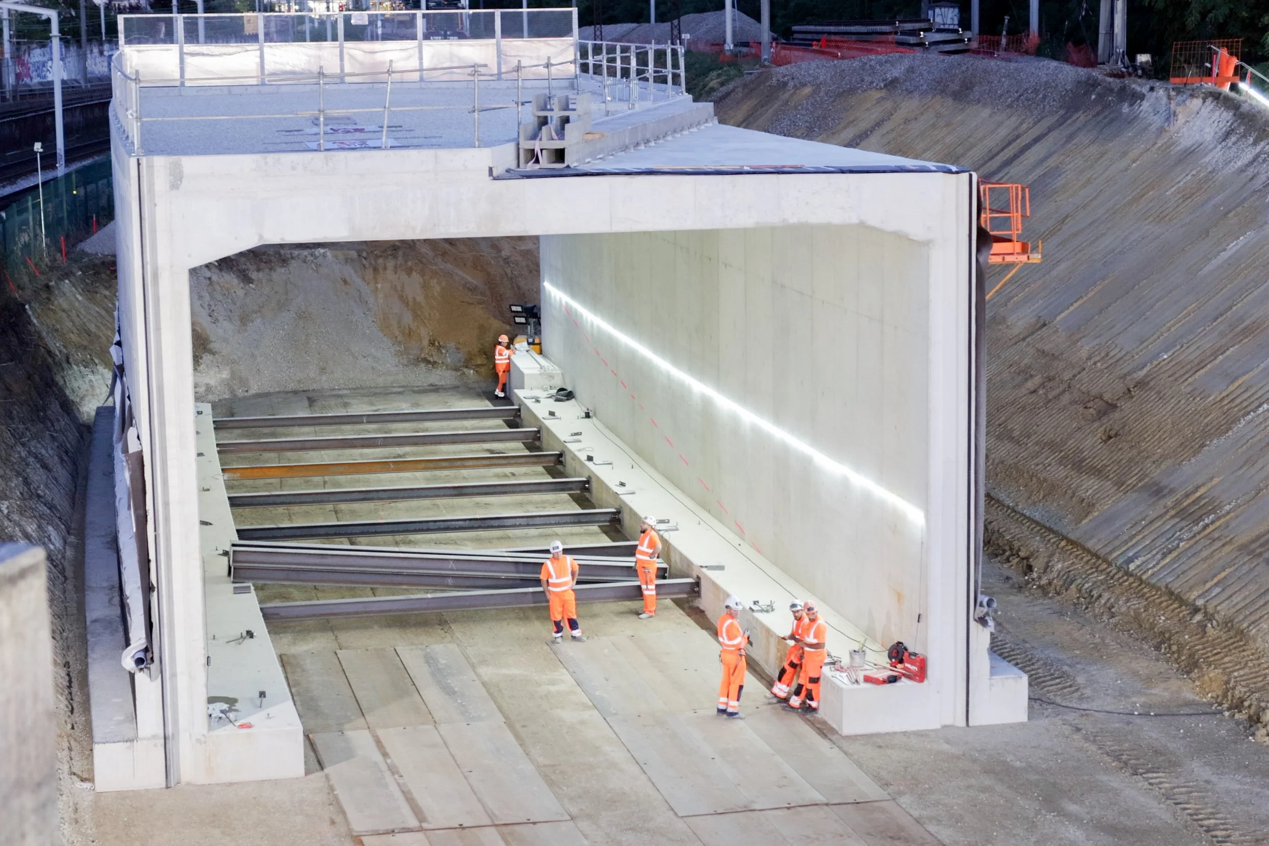 Des ouvriers en vêtements de sécurité orange travaillent sur la construction d'un tunnel ou d'une structure souterraine en béton.”}