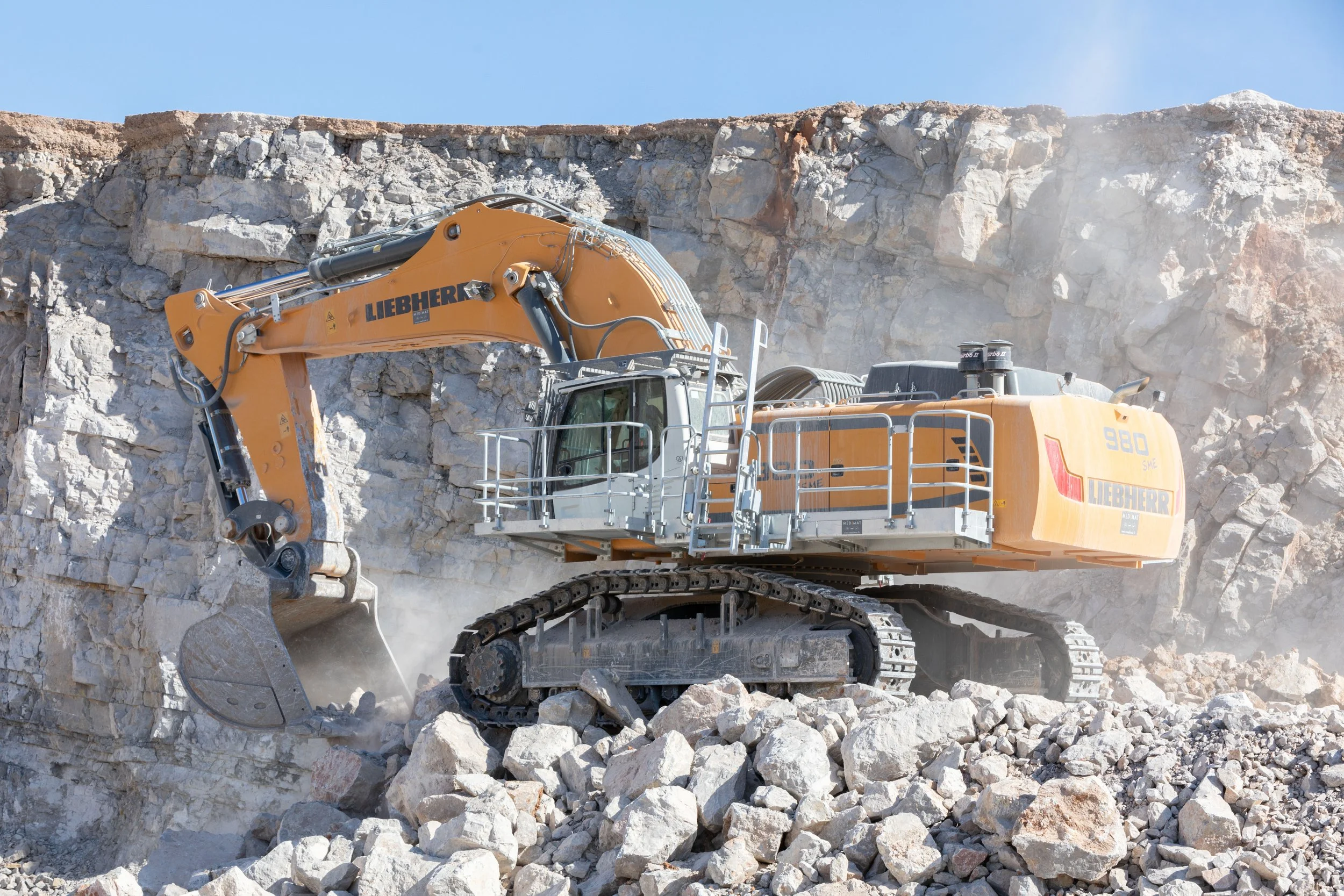Excavatrice jaune de la marque Liebherr en train d'extraire des rochers dans une carrière, avec un mur rocheux en arrière-plan et un ciel bleu.