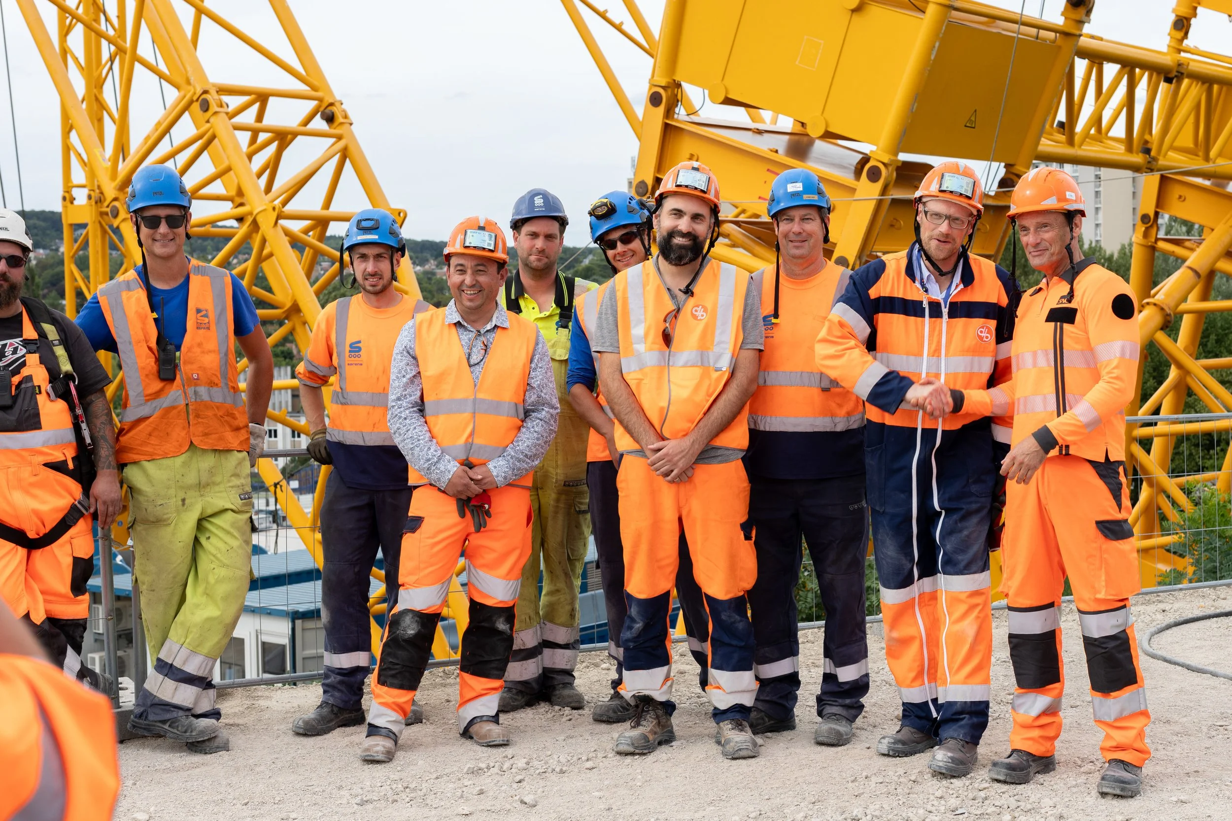 Groupe d'ouvriers de construction portant des casques de sécurité et des gilets de haute visibilité, debout devant une structure en acier jaune.