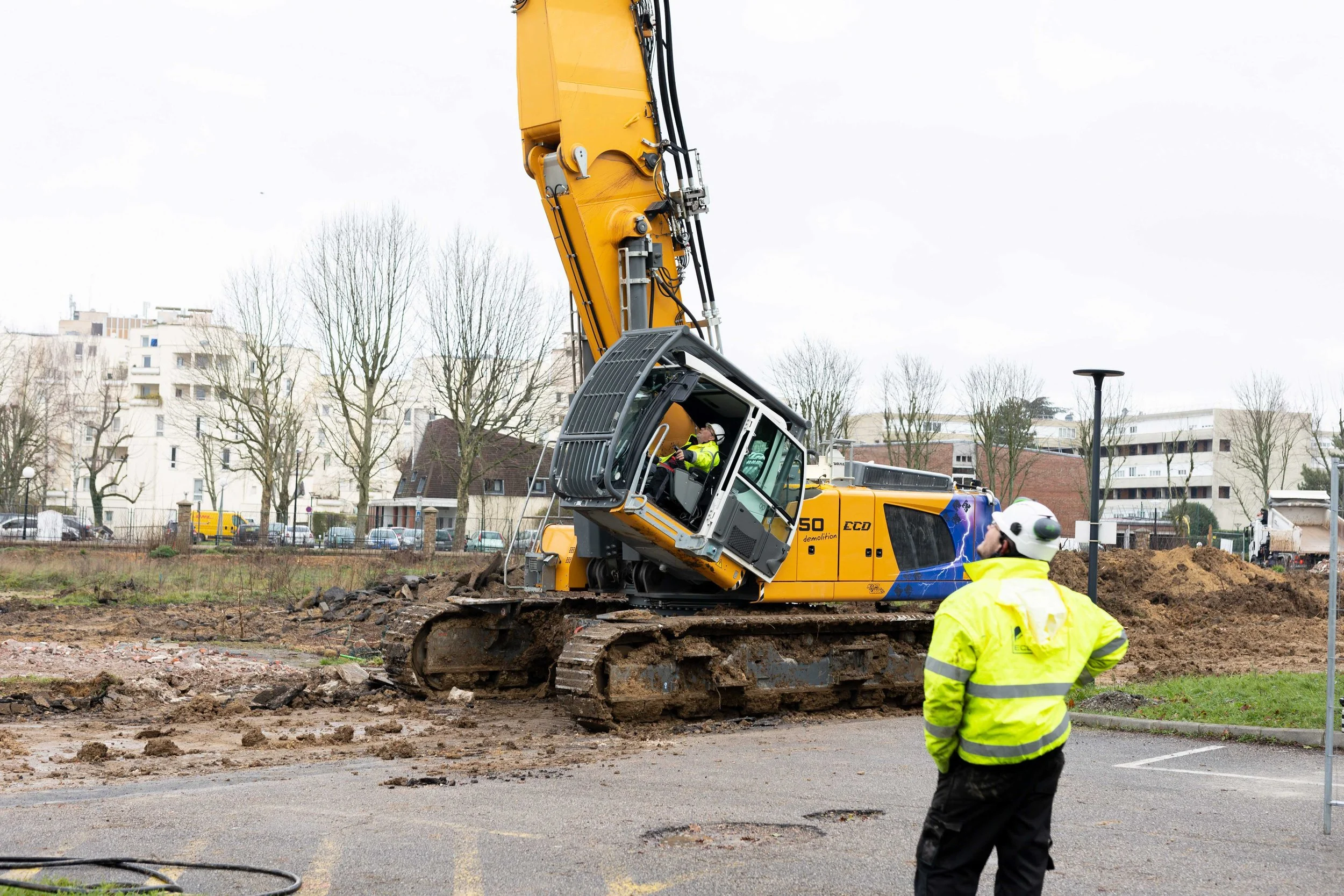 Travail de démolition avec une excavatrice jaune sur un site en extérieur, plusieurs ouvriers en veste jaune et casque de sécurité