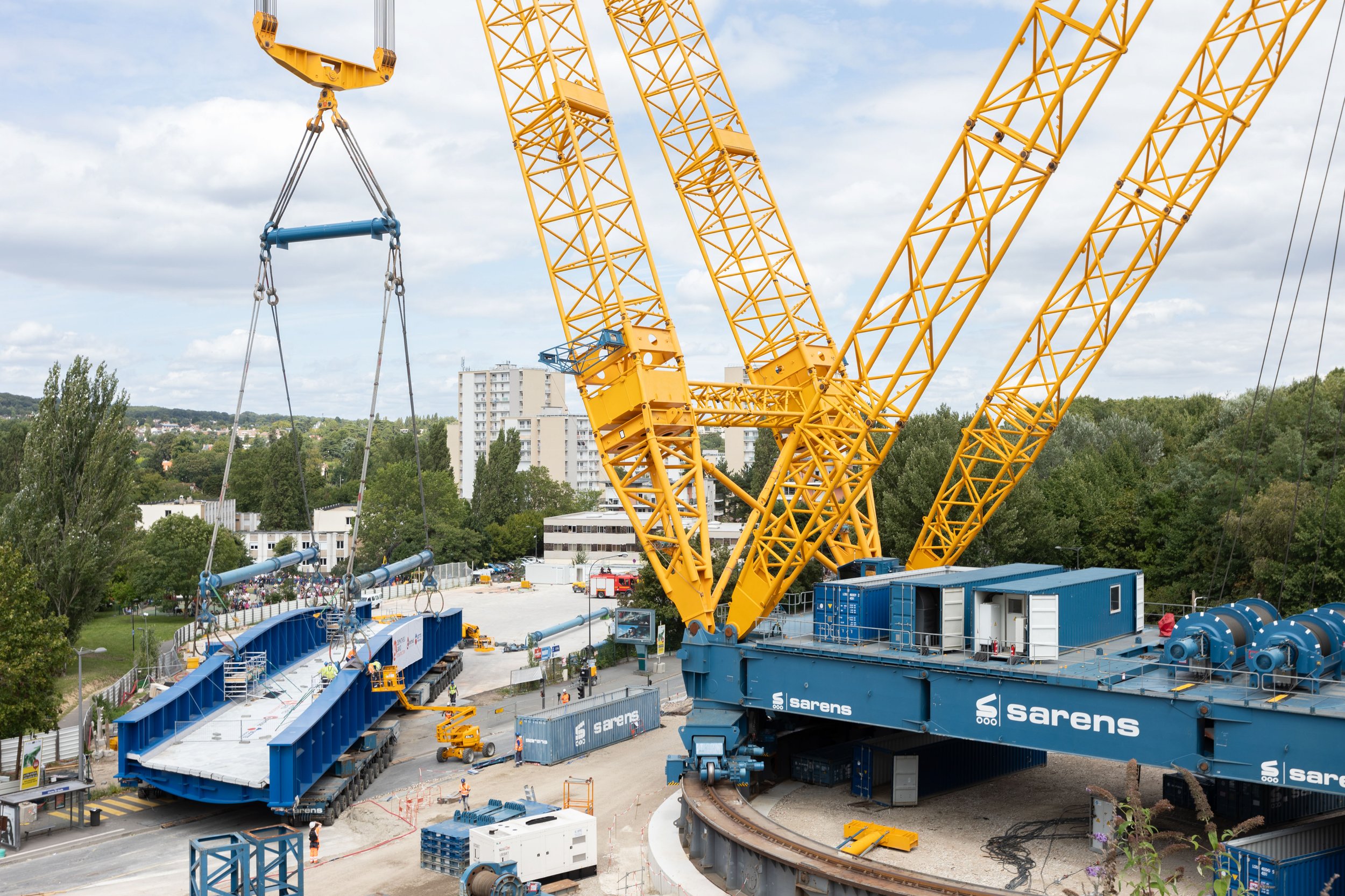 Une grande grue jaune en cours de construction ou d'installation, avec des éléments de structure en acier et un chemin de fer en dessous, dans un environnement urbain avec des bâtiments en arrière-plan.