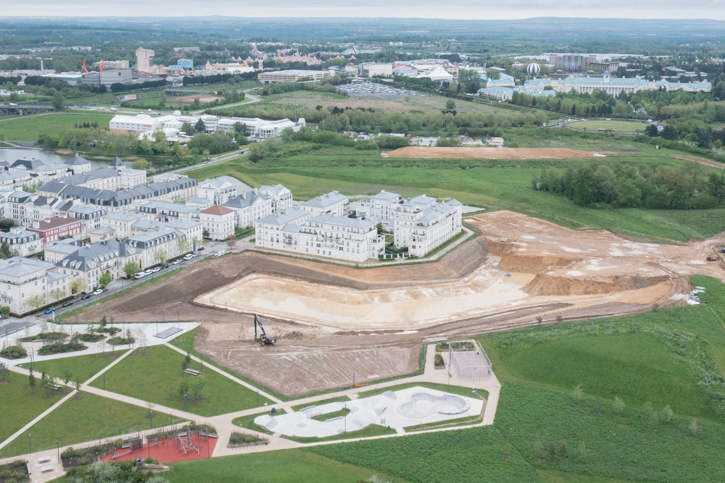 Vue aérienne d'un parc en développement avec des terrains en construction et des bâtiments résidentiels, entouré de zones vertes et d'une ville en arrière-plan.