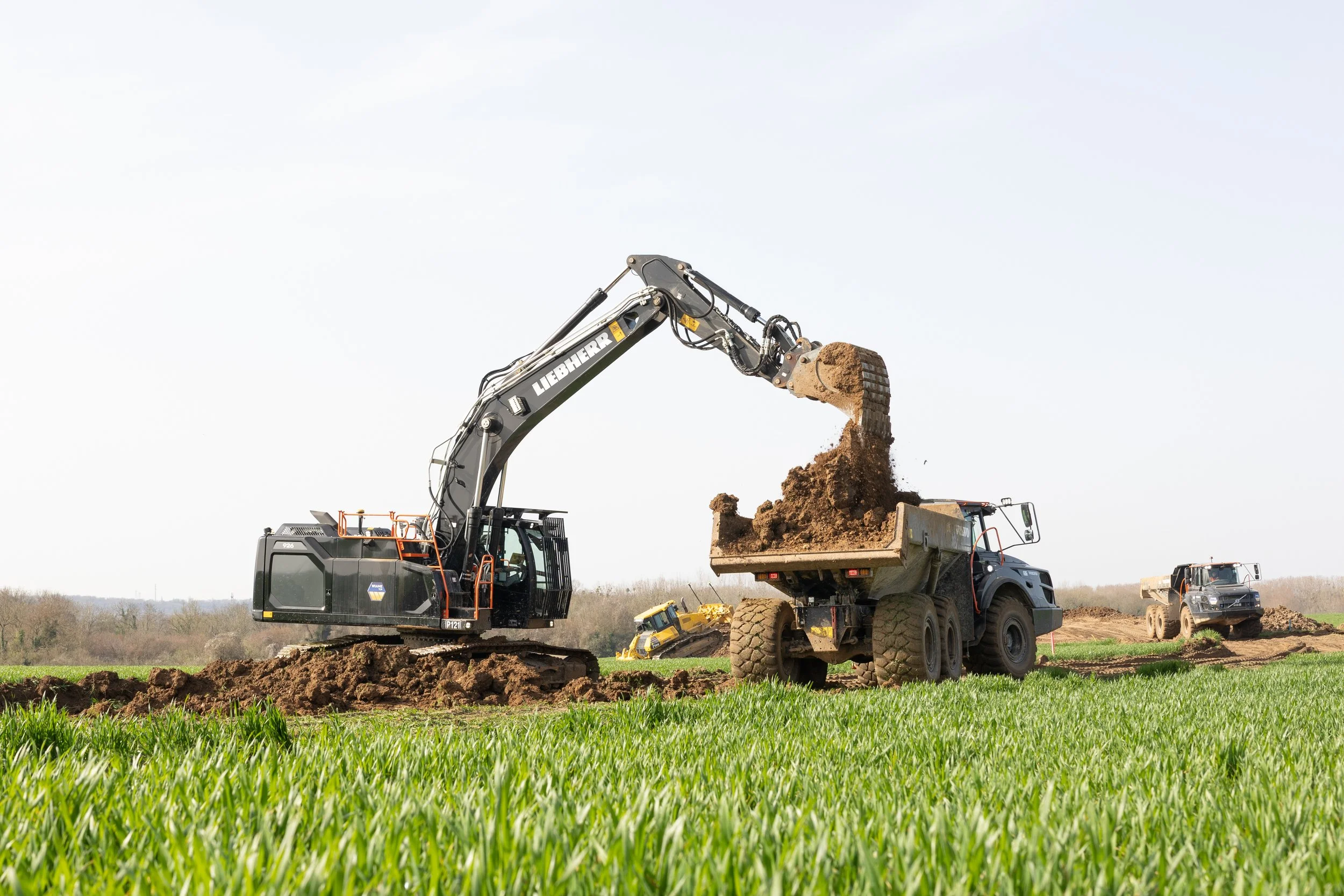 Un excavateur de marque Liebherr décharge de la terre dans une benne sur un chantier, avec un ciel clair en arrière-plan.