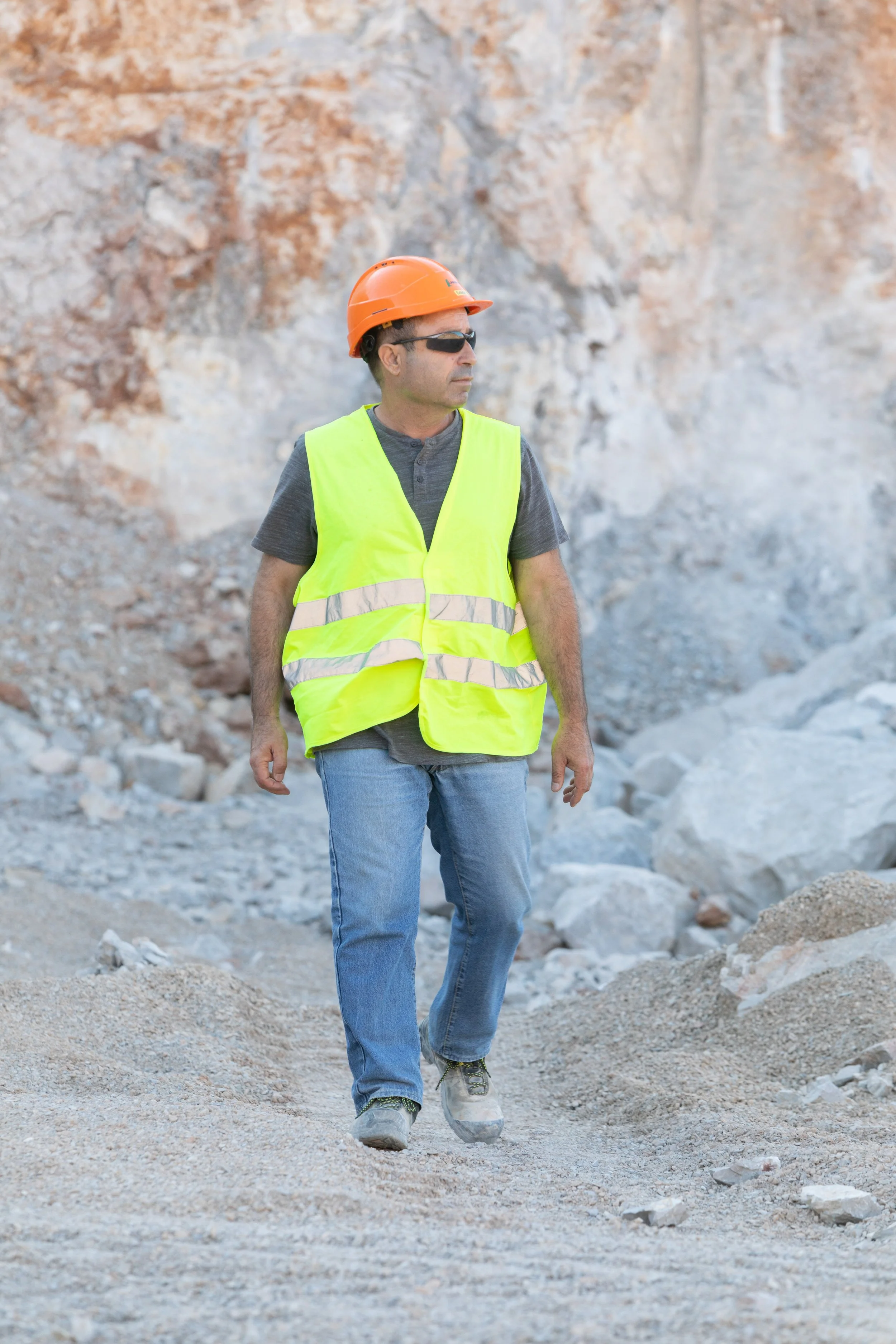 Un homme marchant dans un environnement rocheux, portant un casque orange, des lunettes de soleil, une veste de sécurité jaune fluo, un t-shirt gris, un jean bleu et des chaussures de sécurité.