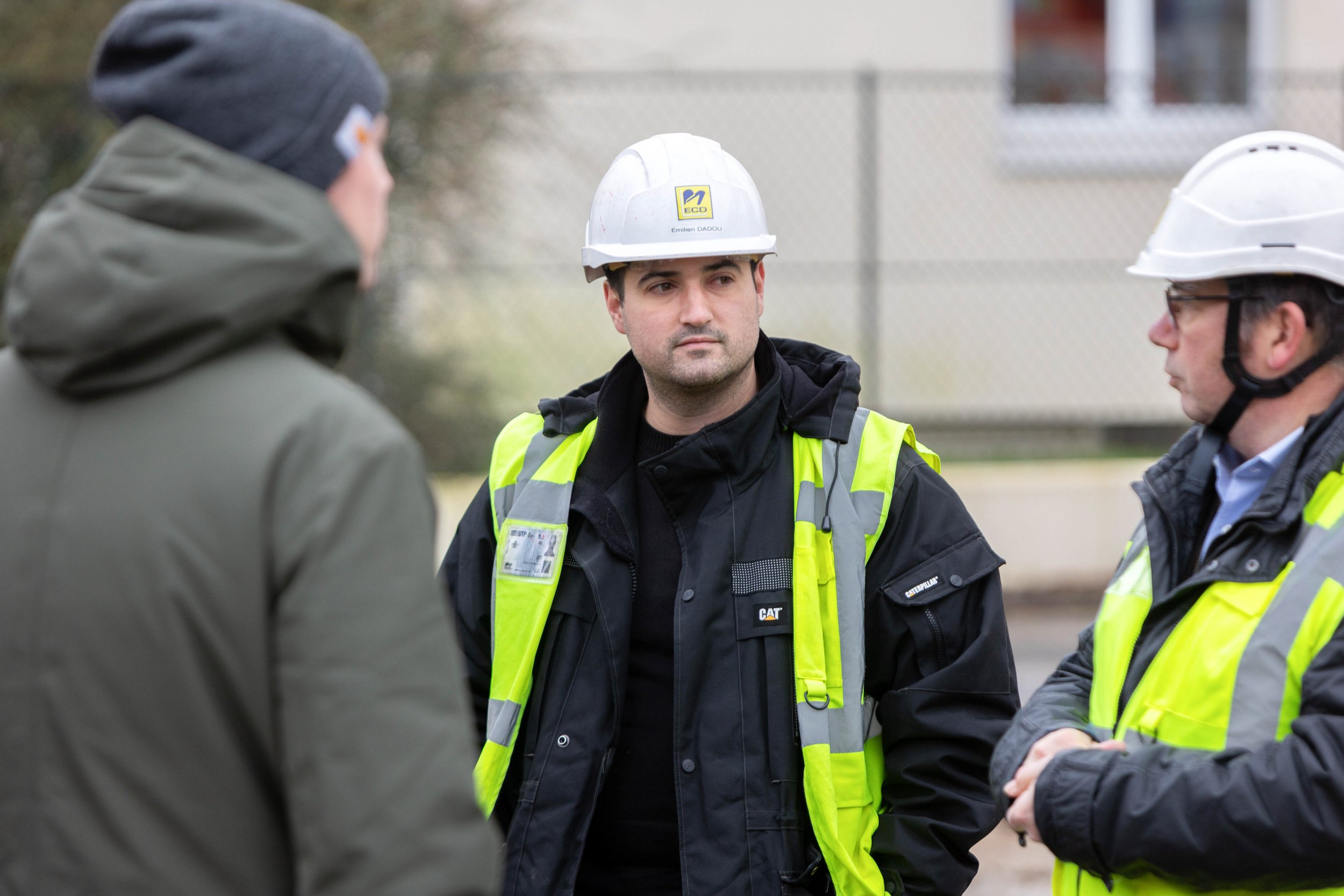 Trois hommes portant des casques de sécurité jaunes avec des vestes de sécurité jaunes discutent à l'extérieur, avec un mur en brique en arrière-plan.