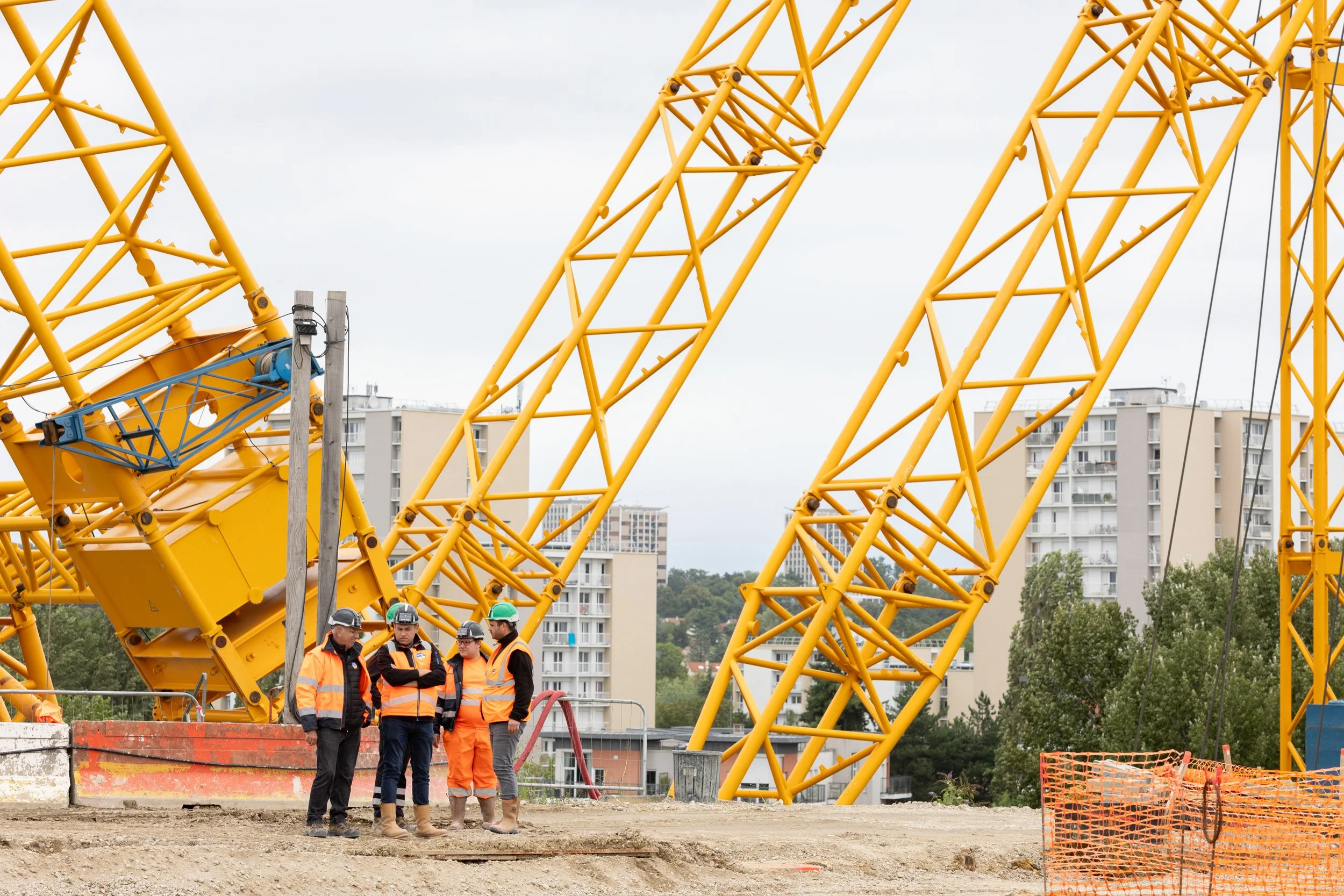 Travailleurs portant des casques et des vêtements de sécurité sur un site de construction avec une grande grue jaune en arrière-plan.