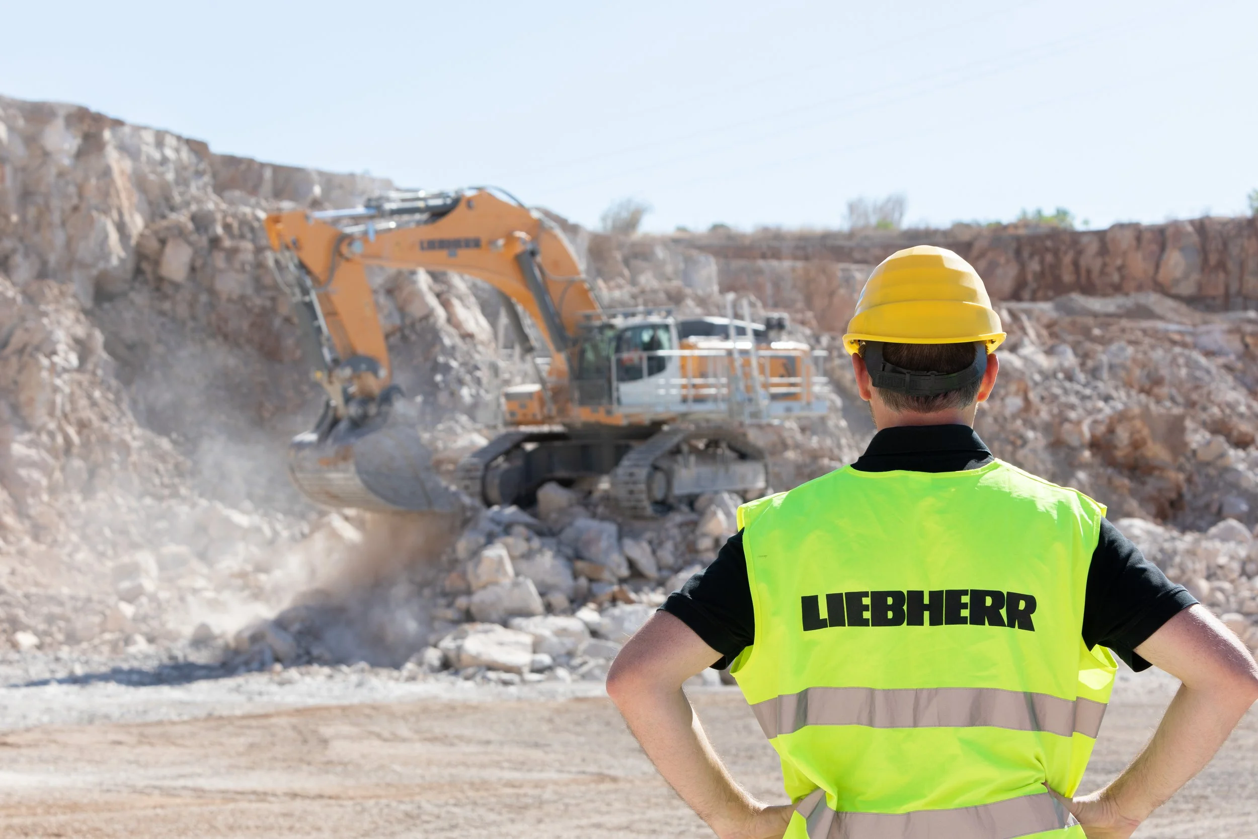 Un homme portant un gilet jaune avec l'inscription "LIEBHERR" à l'arrière observe un chantier de terrassement avec une excavatrice en action, entouré de roches et de fumée