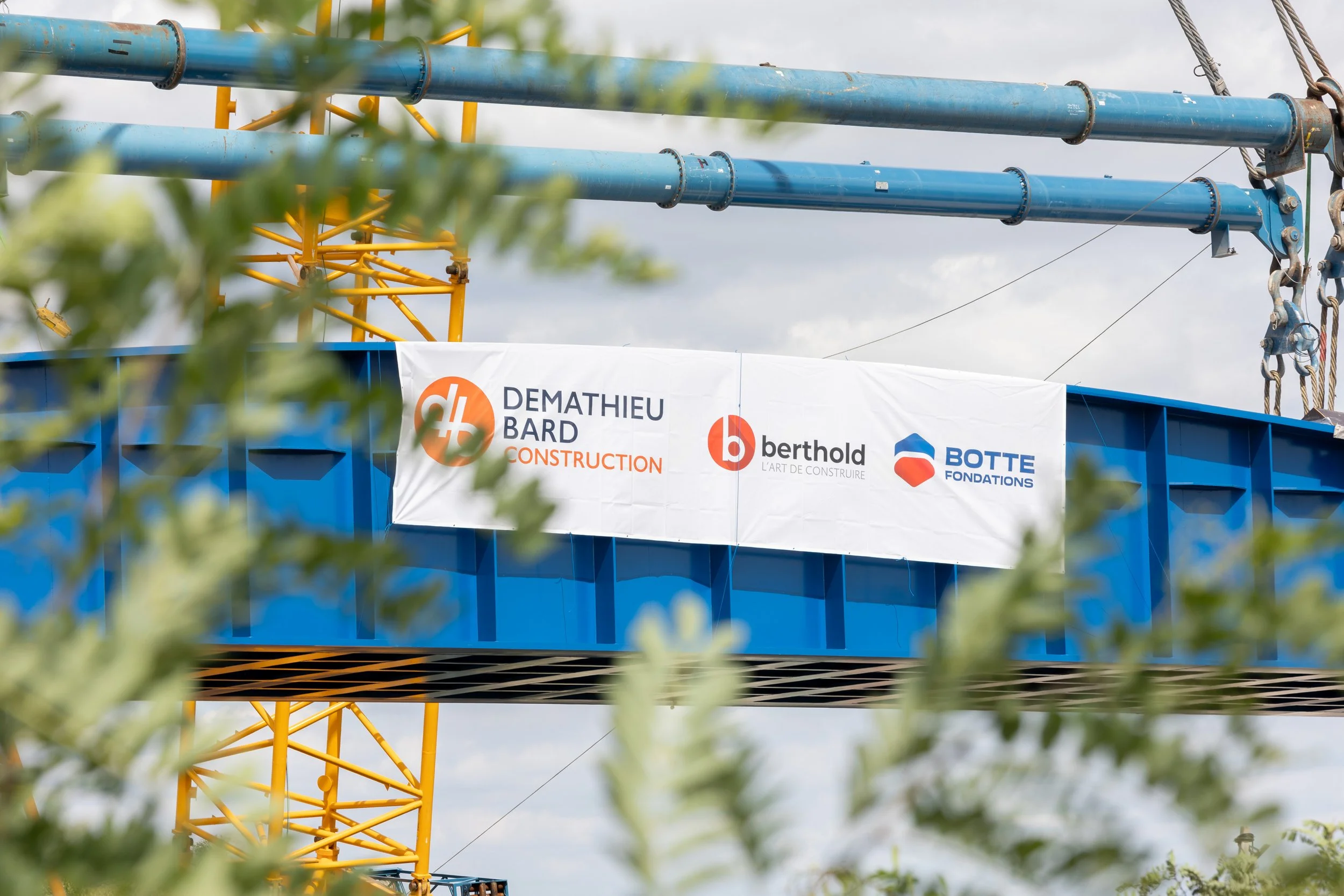 Une structure de construction colorée avec des tuyaux bleus et une tour jaune. Un panneau affiche les logos de Demathieu Bard, berthold et Botte Fondations.