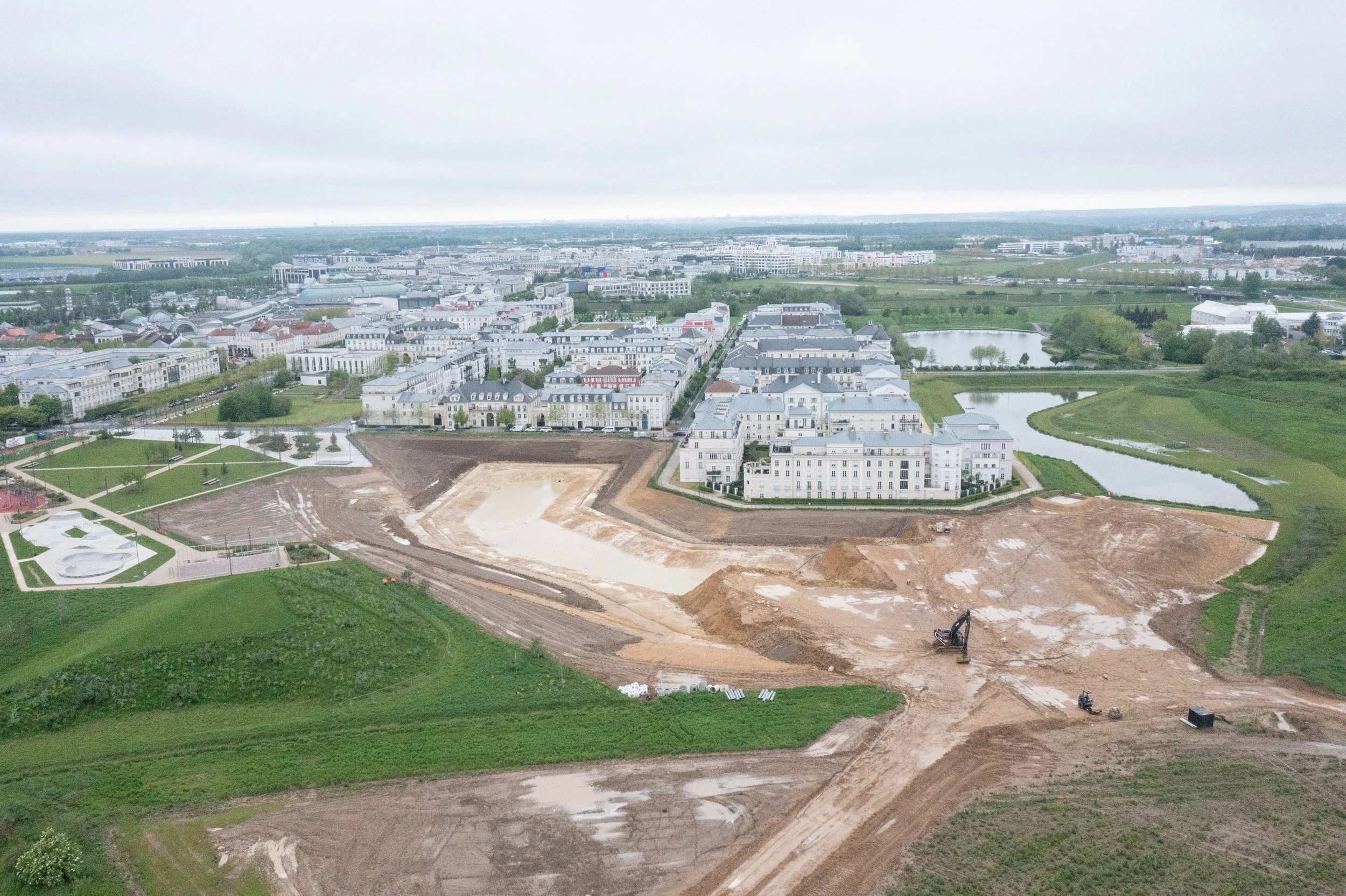 Vue aérienne d'une zone urbaine en développement avec des bâtiments résidentiels, des plans d'aménagement, un lac et des espaces verts, sous un ciel nuageux.