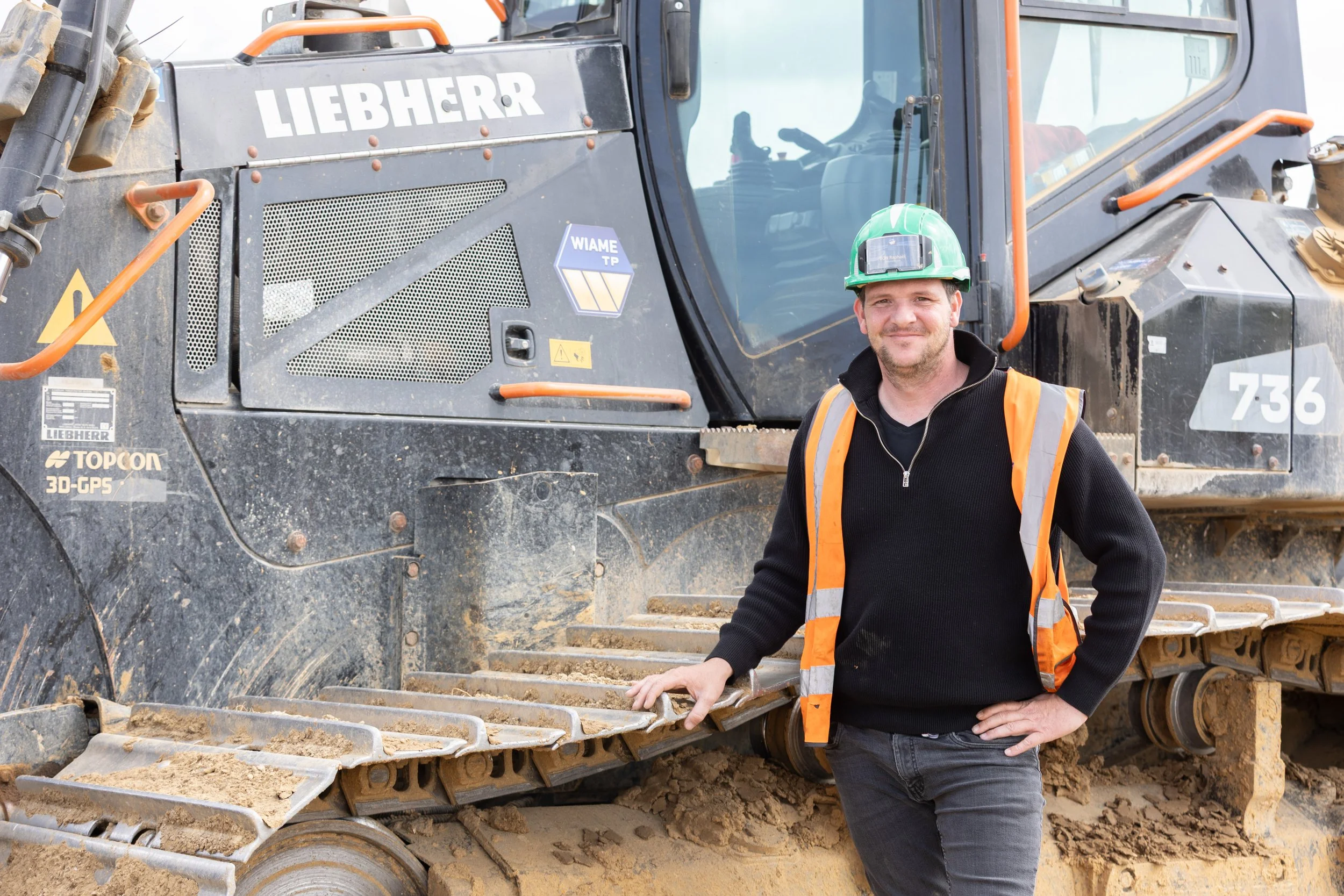 Un homme portant un casque de sécurité vert, un gilet de sécurité orange et un pull noir se tient à côté d'un bulldozer de la marque Liebherr. Il sourit et pose sa main sur le chenillard du véhicule, qui est couvert de terre et de poussière.
