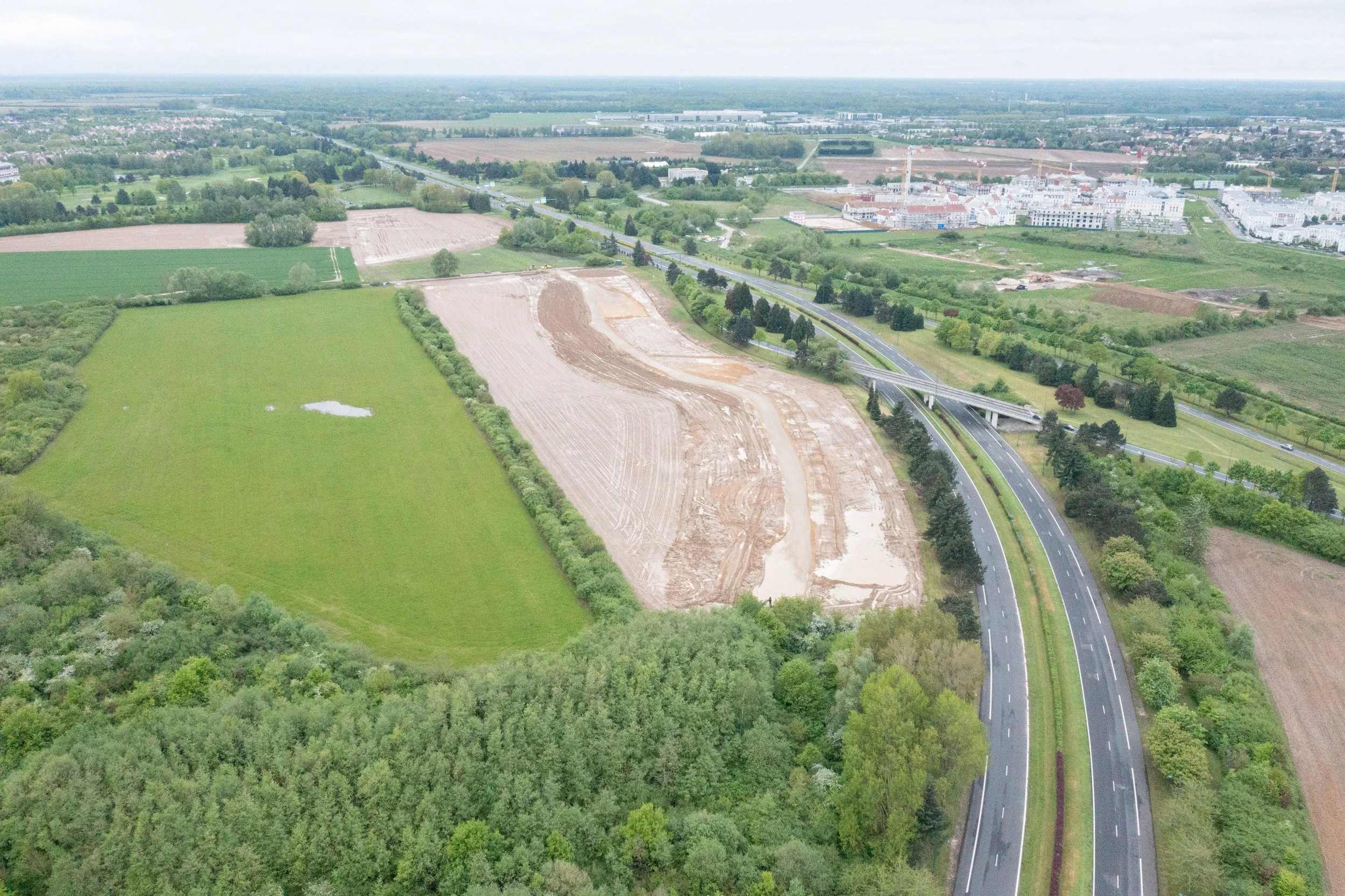Vue aérienne d'une zone en construction avec des parcelles de terre déblayée, une route en courbe et un paysage rural avec champs et forêts et une zone urbaine au fond.