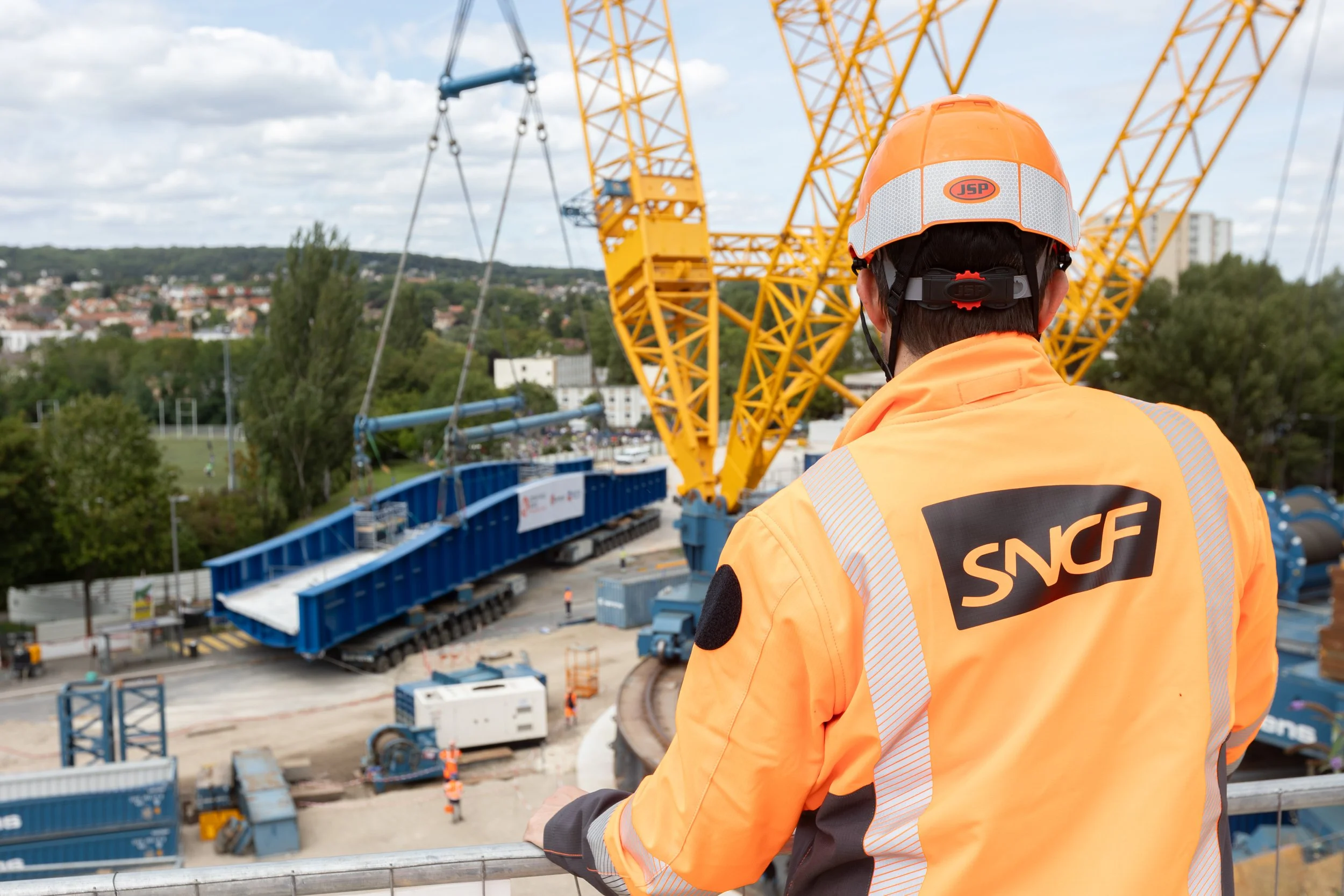 Un ouvrier portant un casque orange et un gilet orange avec la marque SNCF observe un chantier de construction avec une grande grue jaune surplombant la ville en arrière-plan.