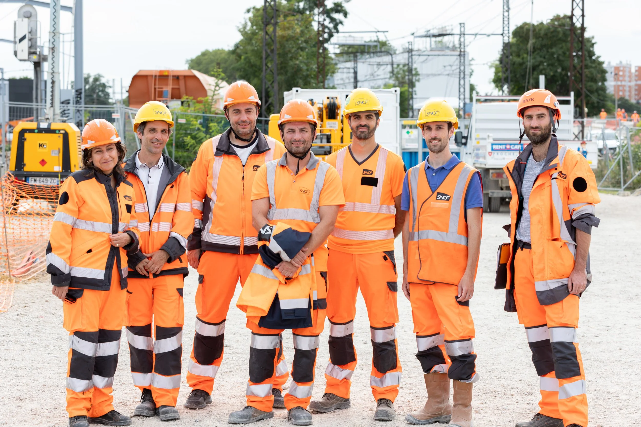 Groupe de travailleurs portant des vêtements de sécurité orange et des casques dans un site de construction ou de maintenance ferroviaire.