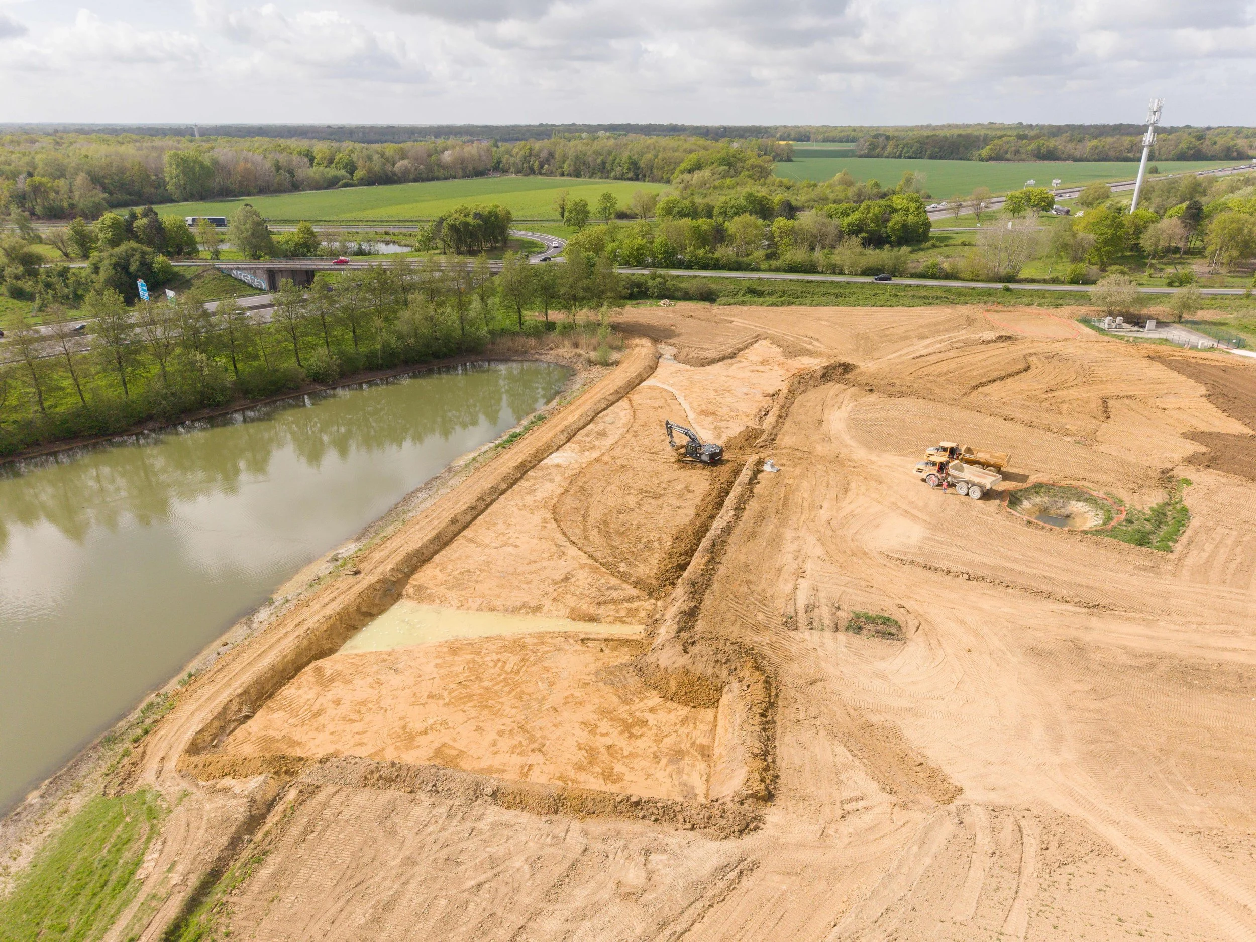 Vue aérienne d'un chantier de construction avec des machines, un étang et une route à proximité, entouré de champs et de forêt sous un ciel nuageux.