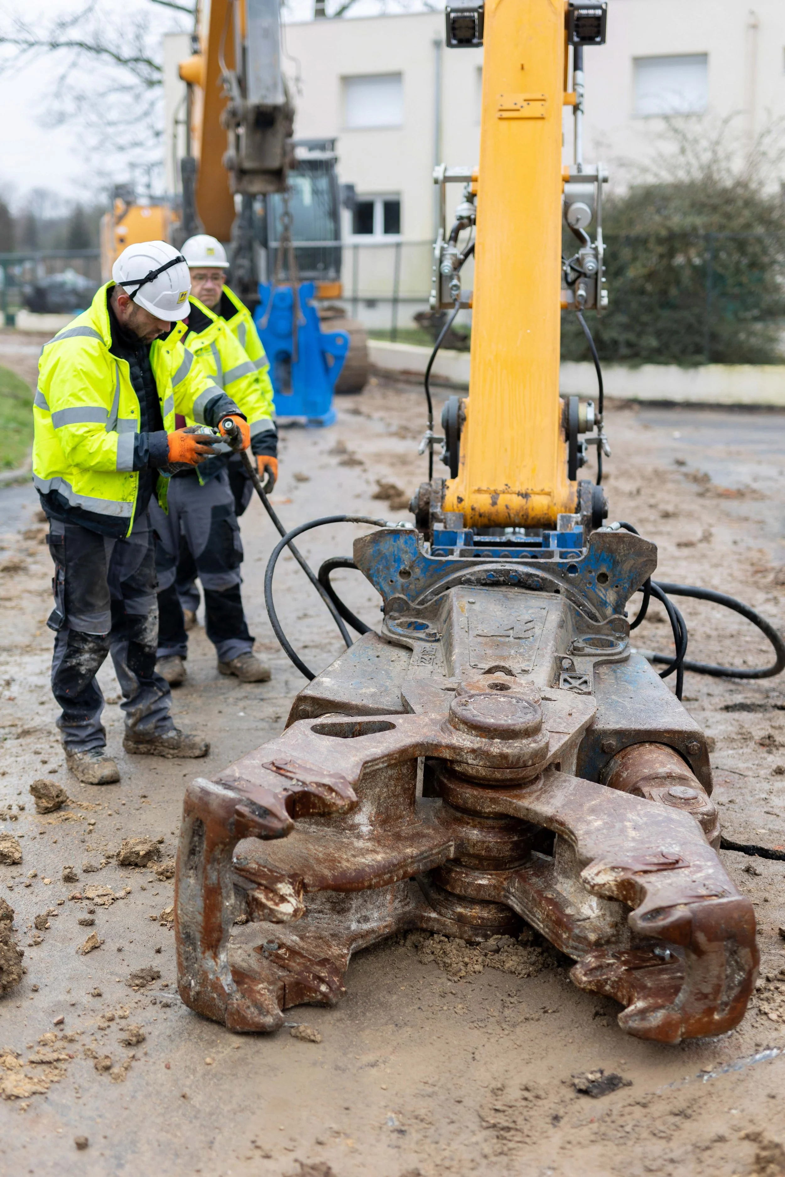 Deux ouvriers en casque et gilet de sécurité travaillent sur une machine de construction avec une pelle hydraulique en arrière-plan. Le sol est en terre et délimité par un chantier urbain.