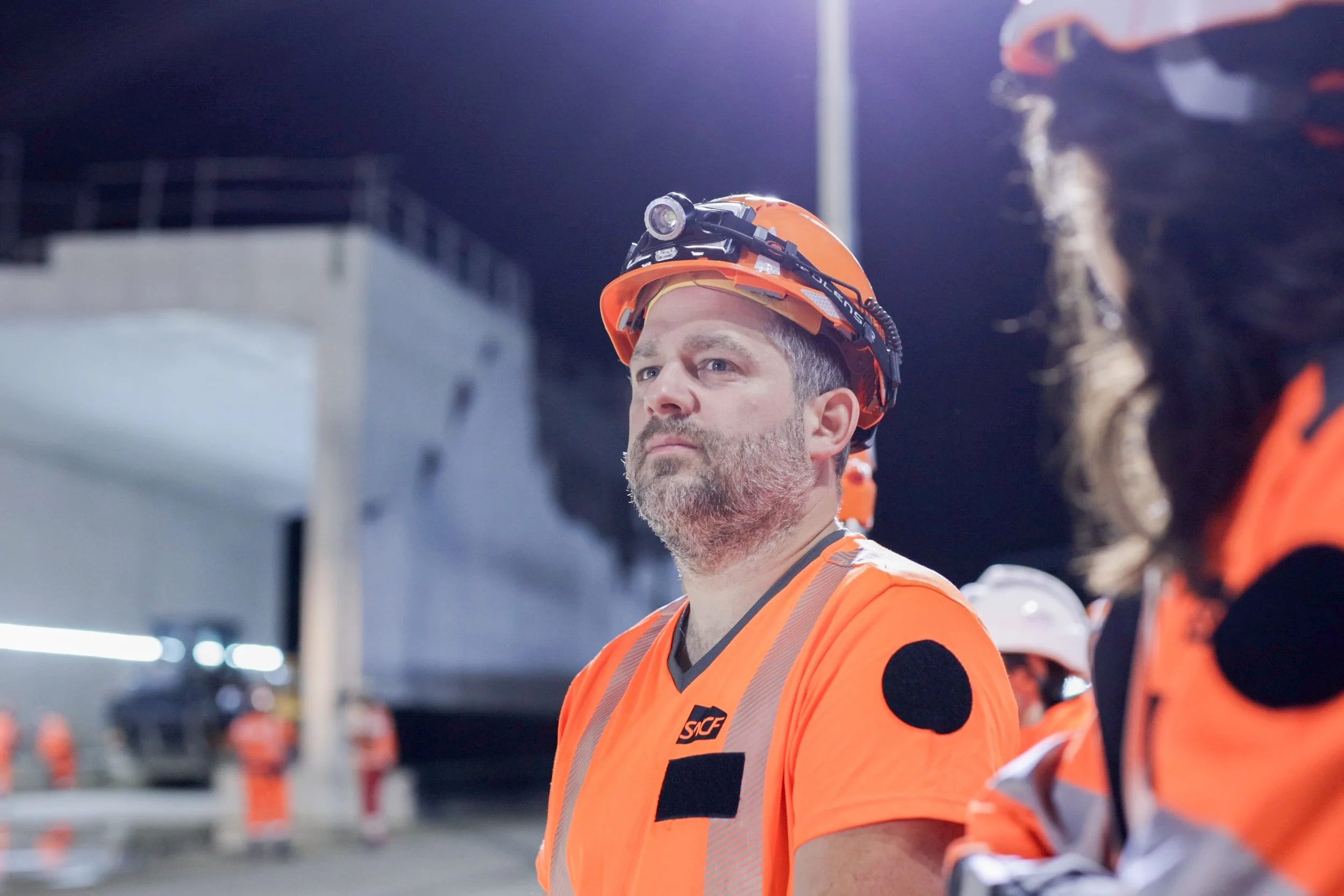 Un homme portant un casque de sécurité orange et un gilet de sécurité orange en groupe, probablement lors d'une inspection ou d'une opération dans une zone industrielle ou un site de construction, la scène se déroule la nuit.