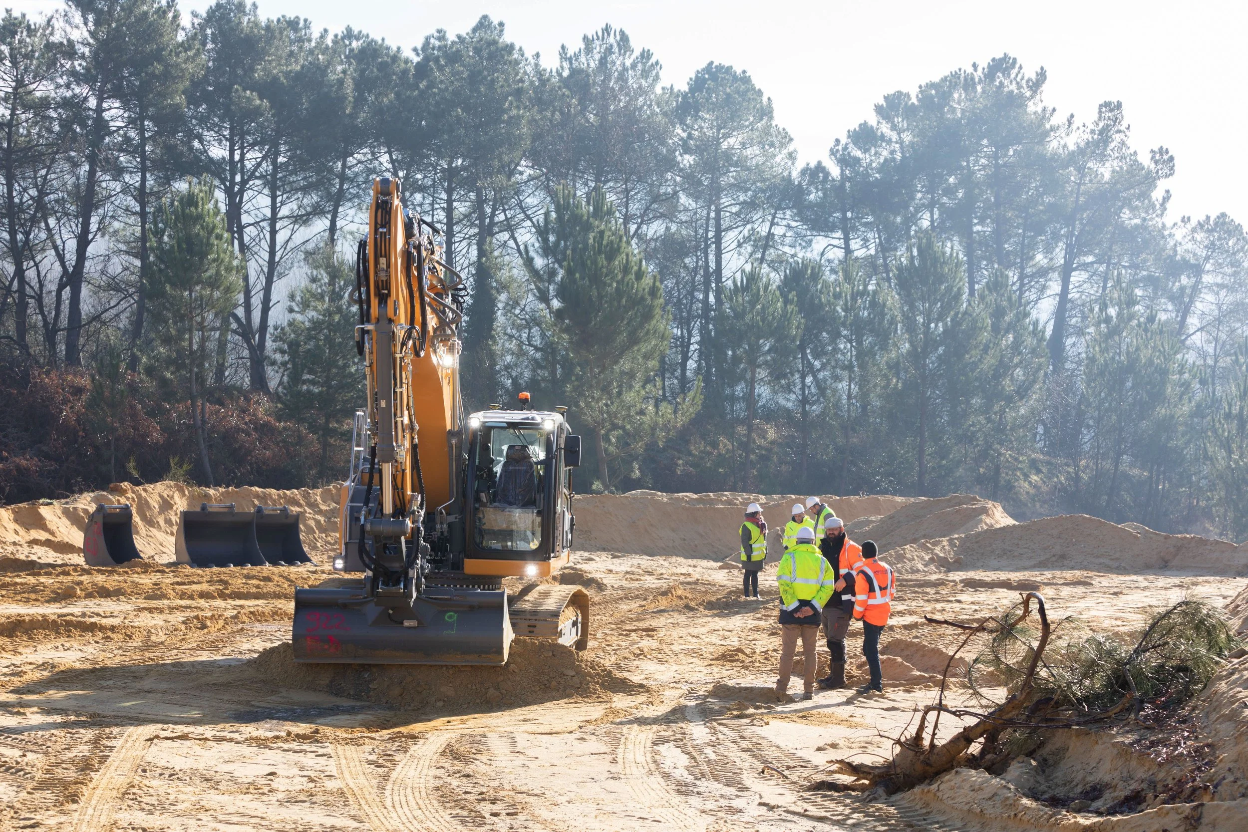 Travailleurs en sécurité vestes jaunes et casques blancs dans un chantier forestier avec un excavateur jaune et des arbres en arrière-plan.