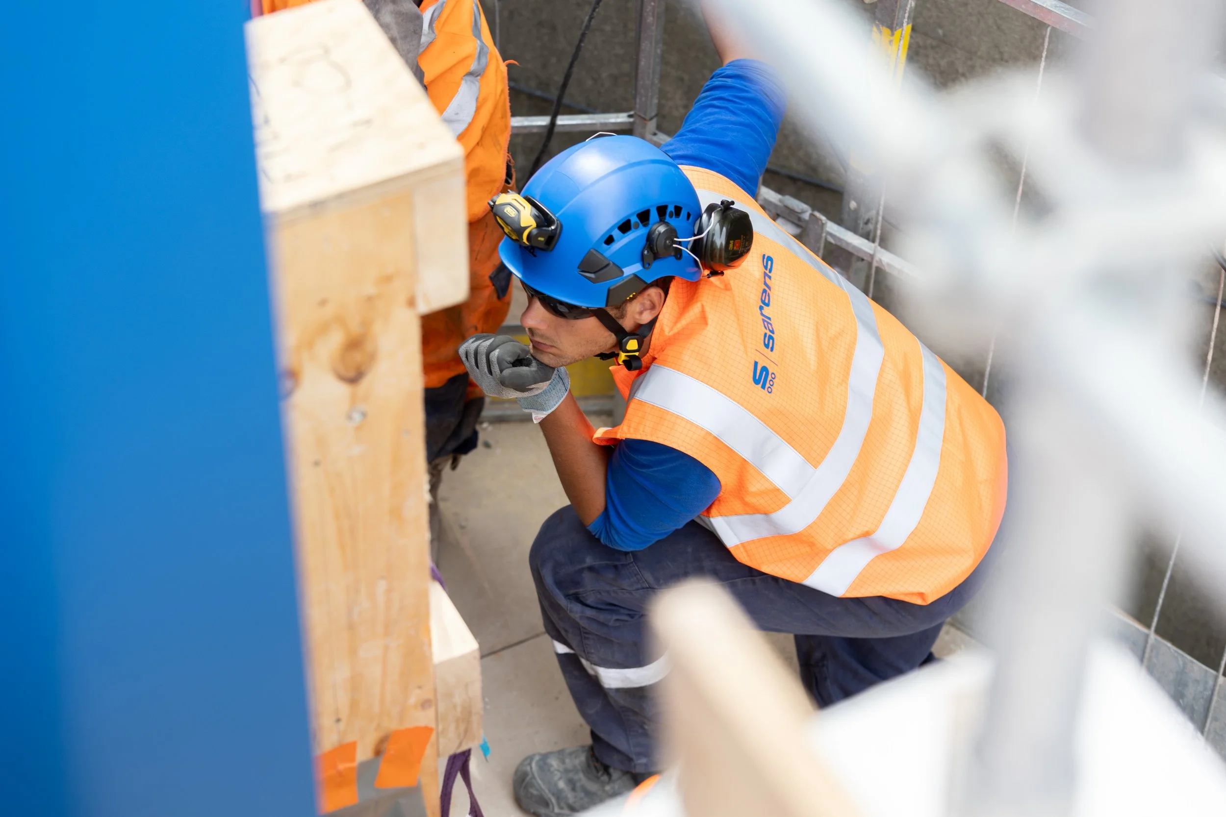 Un ouvrier en casque bleu, gilet orange et lunettes de protection travaille à une construction, penché sur un mur en béton.