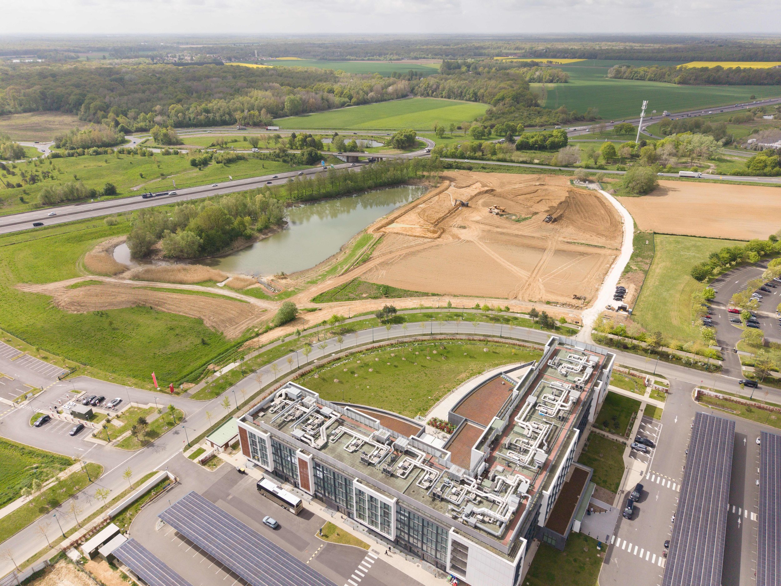 Vue aérienne d'un immeuble moderne avec panneaux solaires, avec des parkings, des routes, une végétation abondante, un étang, et des champs agricoles au loin, sous un ciel partiellement nuageux.
