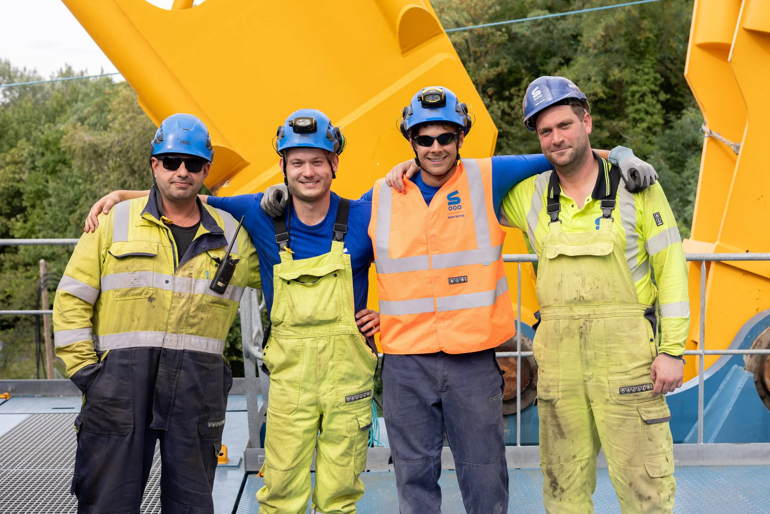 Quatre ouvriers portant des casques bleus, des lunettes de soleil et des vestes de sécurité orange ou jaunes, posant pour une photo devant un équipement de chantier, avec de la végétation en arrière-plan.
