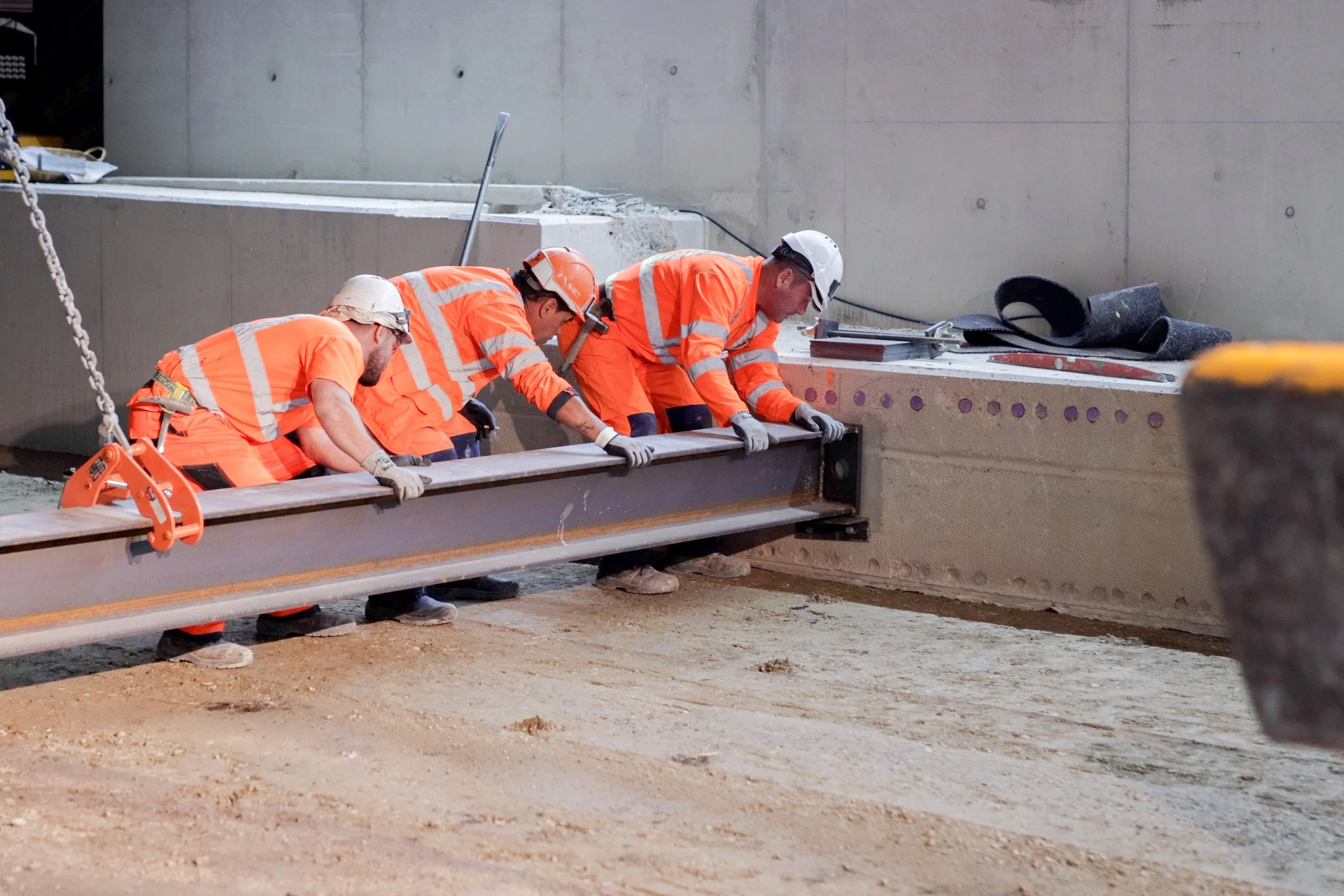 Trois ouvriers en sécurité travaillant sur une structure en construction, portant des vêtements orange et des casques de sécurité, dans un environnement de chantier.