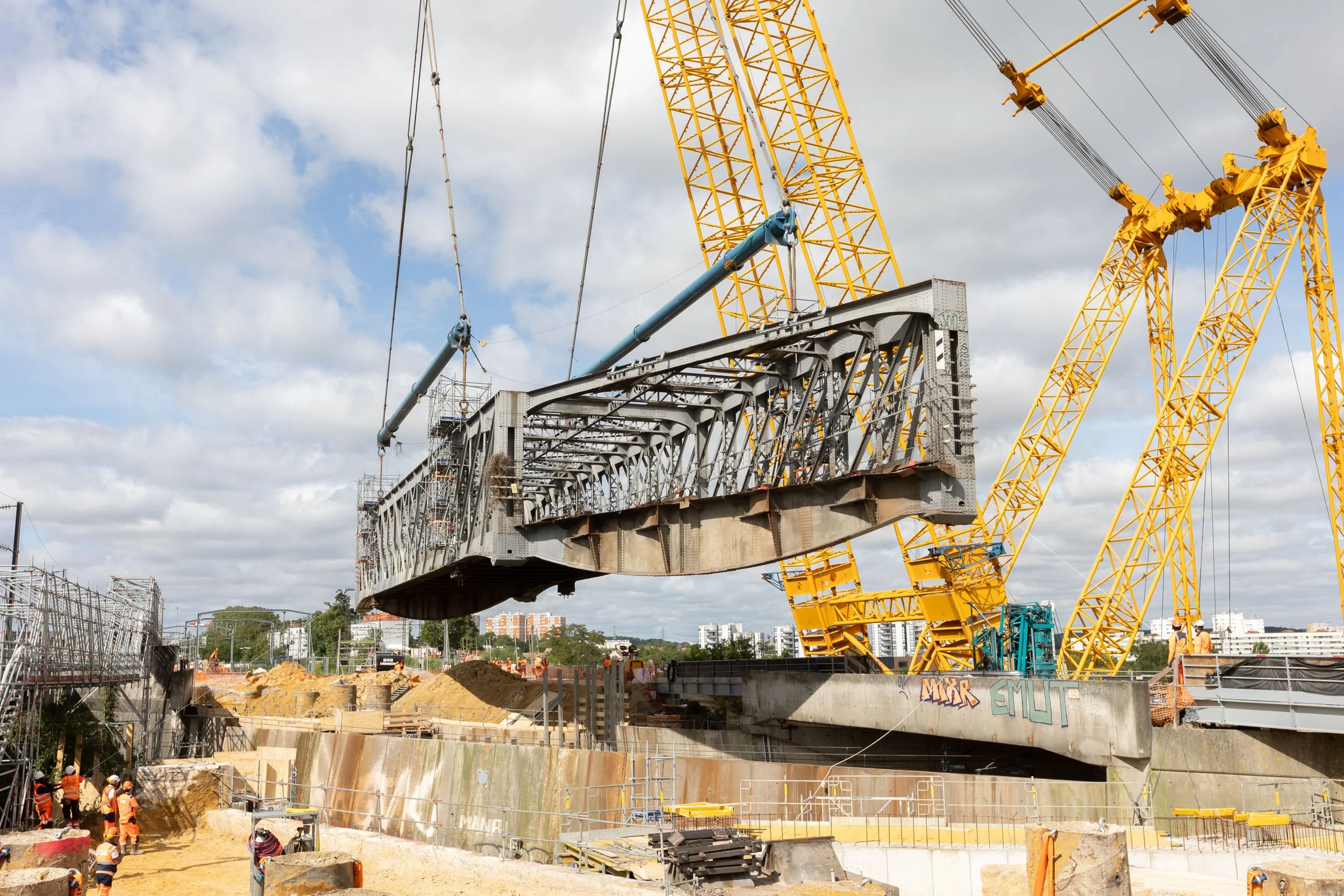 Construction de pont avec grues jaunes et structures métalliques, ouvriers sur le site, ciel nuageux.