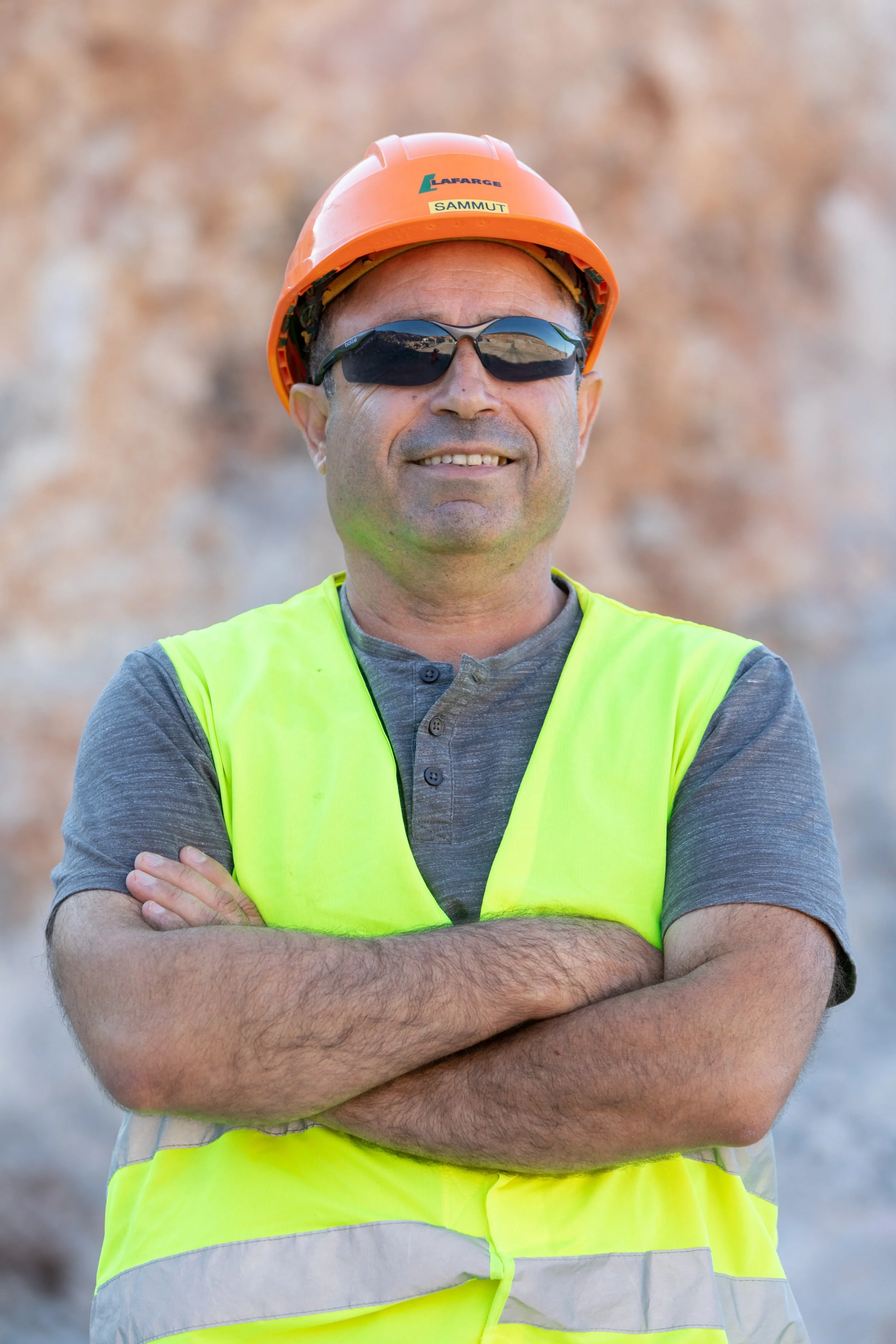 Un homme portant un casque orange, des lunettes de soleil noires, un gilet jaune de sécurité et une chemise grise, croise les bras, vers un fond rocheux.