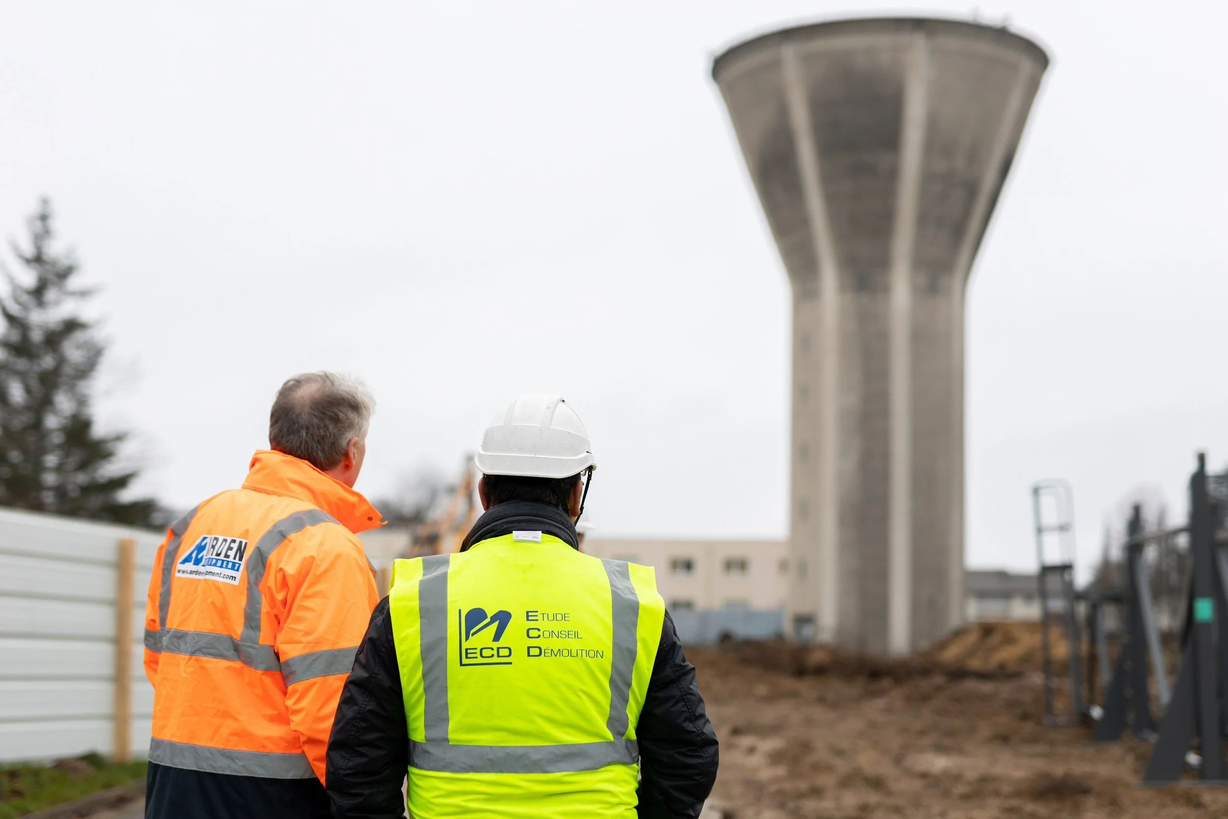 Trois travailleurs portant des gilets de sécurité, deux en orange et un en jaune, regardant un grand réservoir ou une tour de contrôle en béton dans un site de construction ou d'infrastructure.