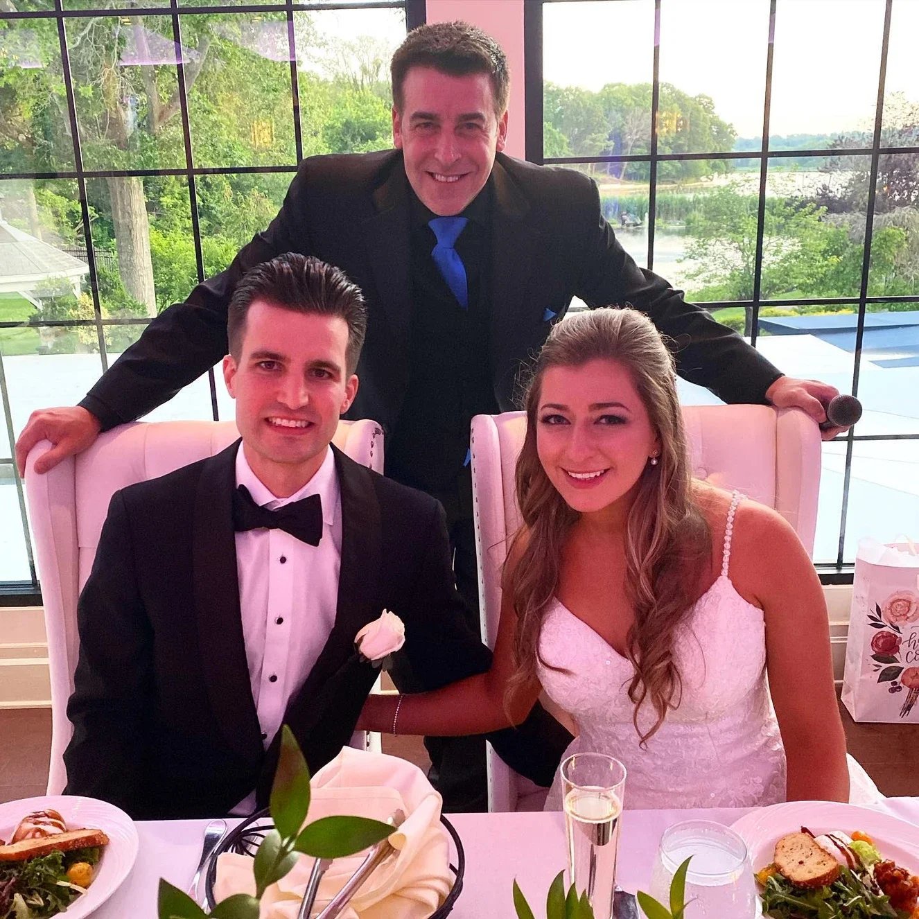 A smiling man in a tuxedo with a bowtie and a woman in a white wedding dress sitting at a table, with another man standing behind them, inside a large glass windowed room with greenery in the background.