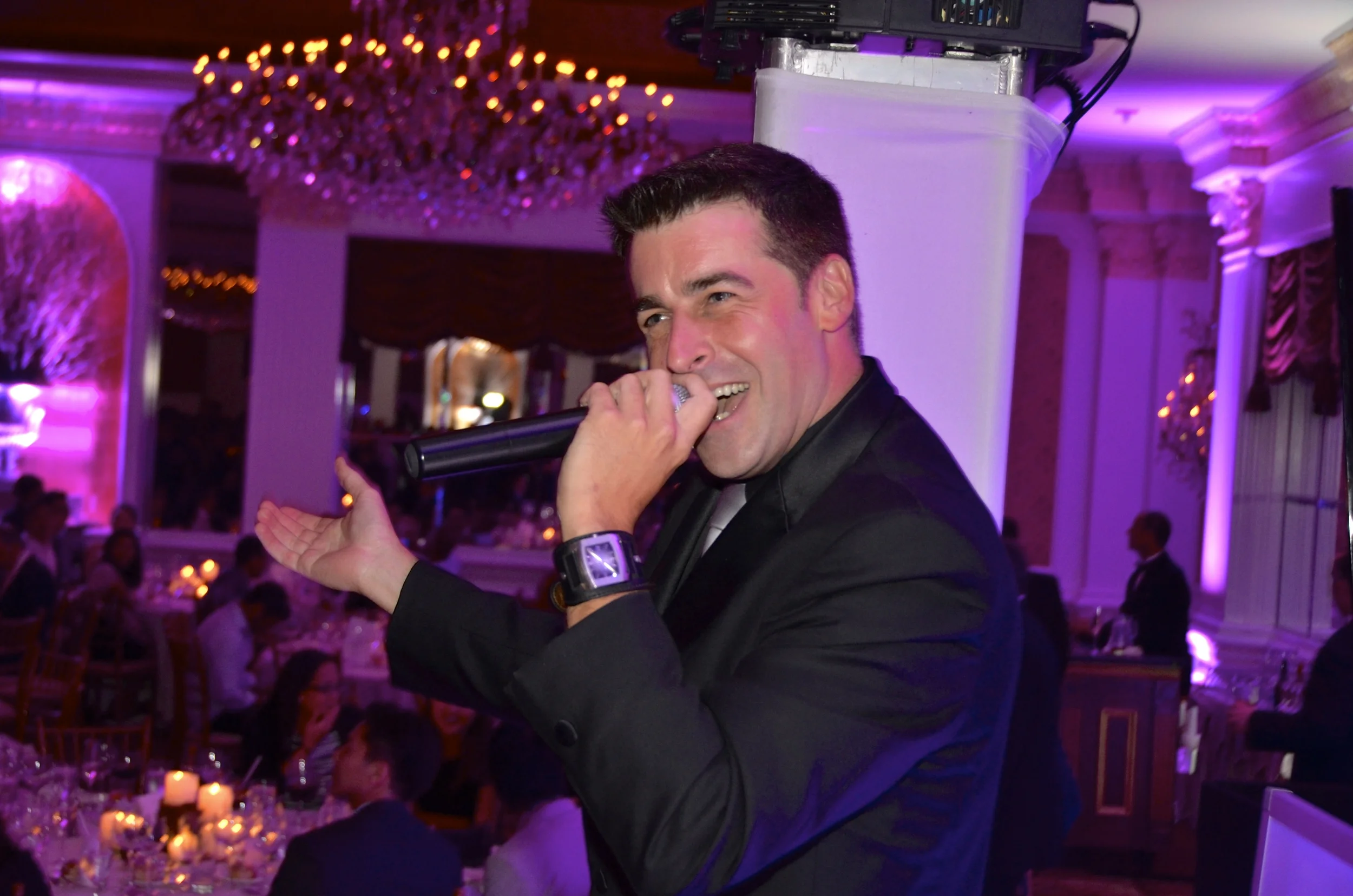 A man in a black jacket holding a microphone, smiling, and performing at an event with purple lighting and elegant decor.