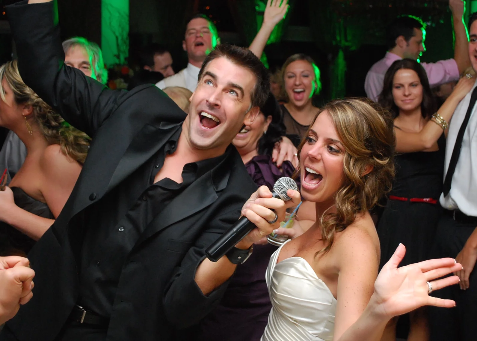 People at a lively celebration, with a man and woman singing into a microphone, surrounded by others dancing and having fun.