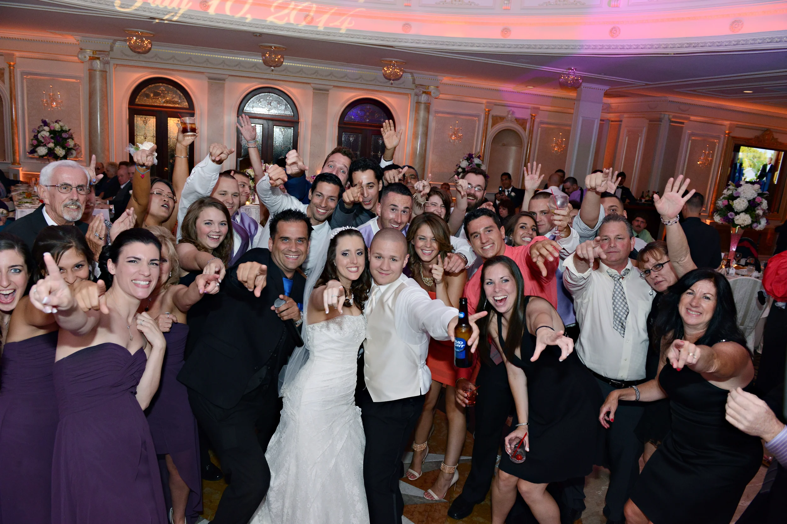A large group of people celebrating at a wedding reception, with the bride and groom in the center, all smiling and posing for the photo in an elegant banquet hall.